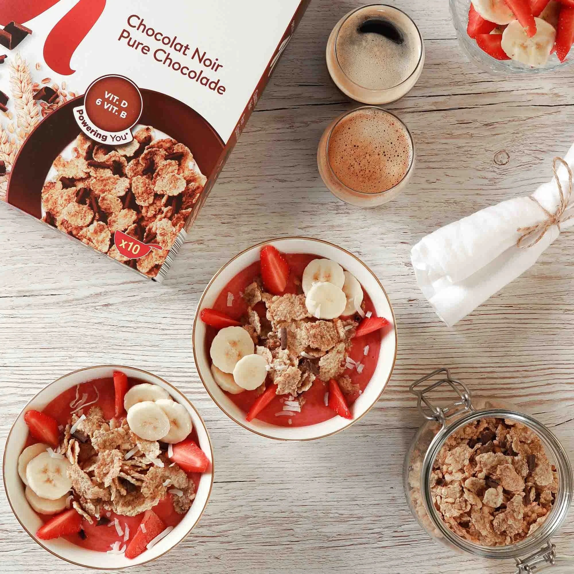 Céréales au chocolat servies avec des tranches de banane et des morceaux de fraise, accompagnées de deux tasses de café et d'une boite de céréales