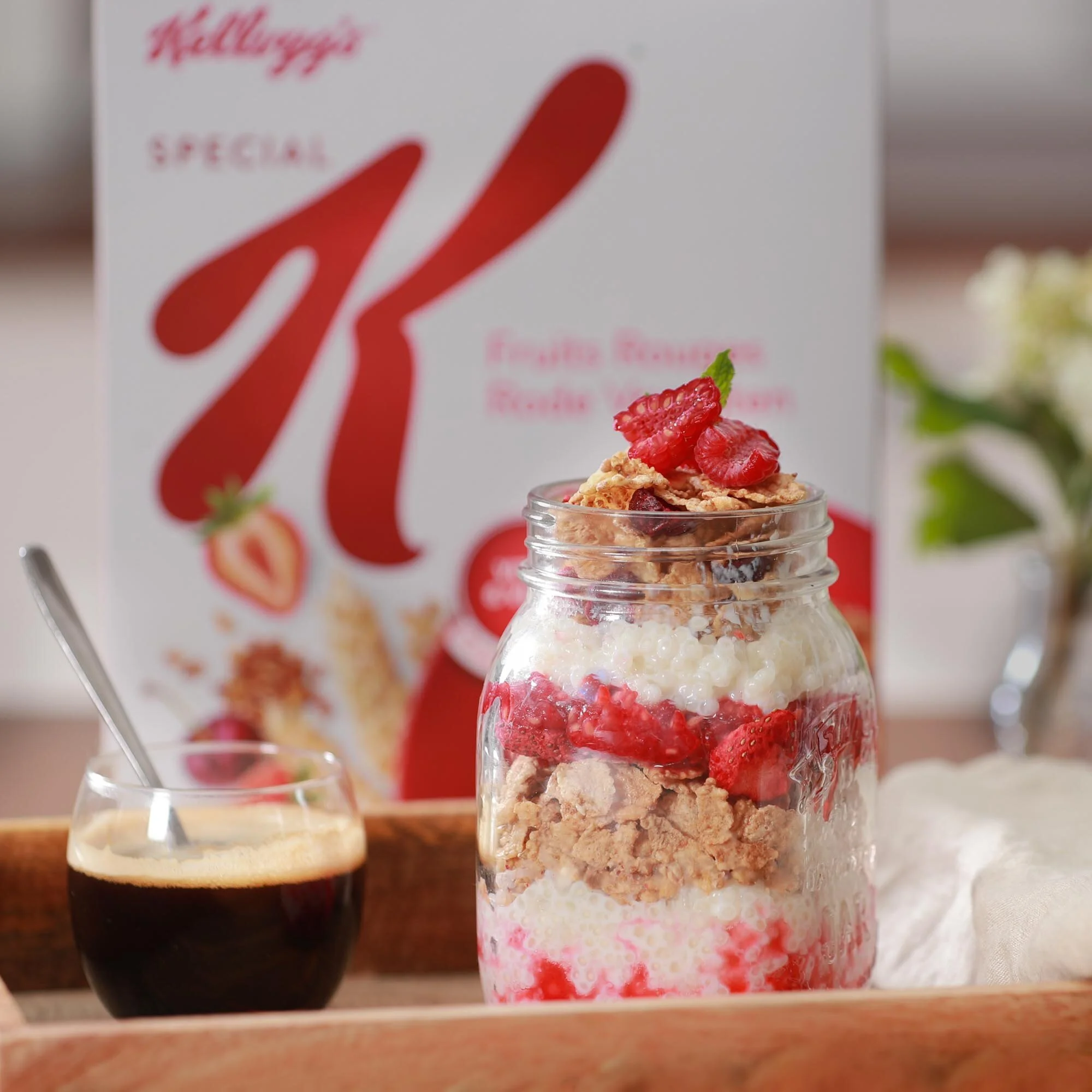 Un pot en verre transparent rempli de couches de céréales, fraises, et lait, avec des morceaux de céréales et des fraises en garniture, placé à côté d'un petit verre d'espresso. En arrière-plan, une boîte de céréales Kellogg's Special K avec une illu
