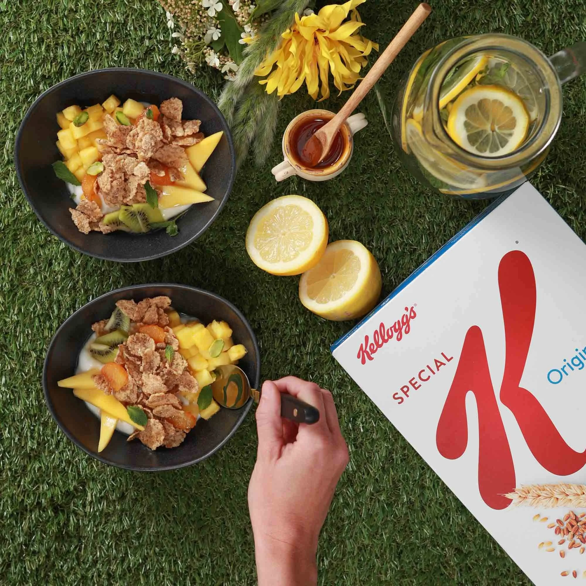Deux bols de fruits frais avec des morceaux de kiwi, de mangue et des céréales, une boîte de céréales Kellogg's Special K, une bouteille d'eau avec des rondelles de citron, deux citrons coupés en deux, une tasse de sirop ou de miel, une main tenant u