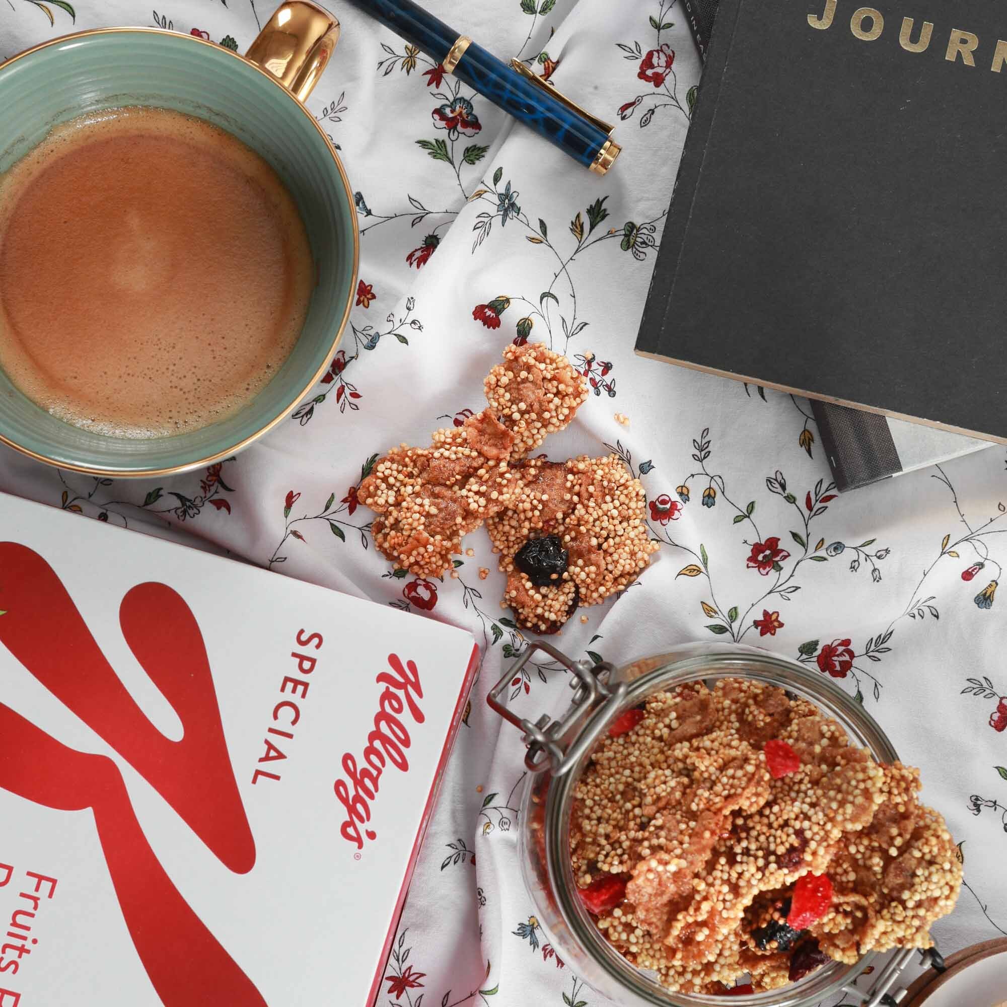 Pause-café avec une tasse de café au lait, desserts croustillants au muelsi et fruits, une boîte Frostory, un cahier noir, un stylo bleu, et un livre noir sur une nappe à motifs floraux.