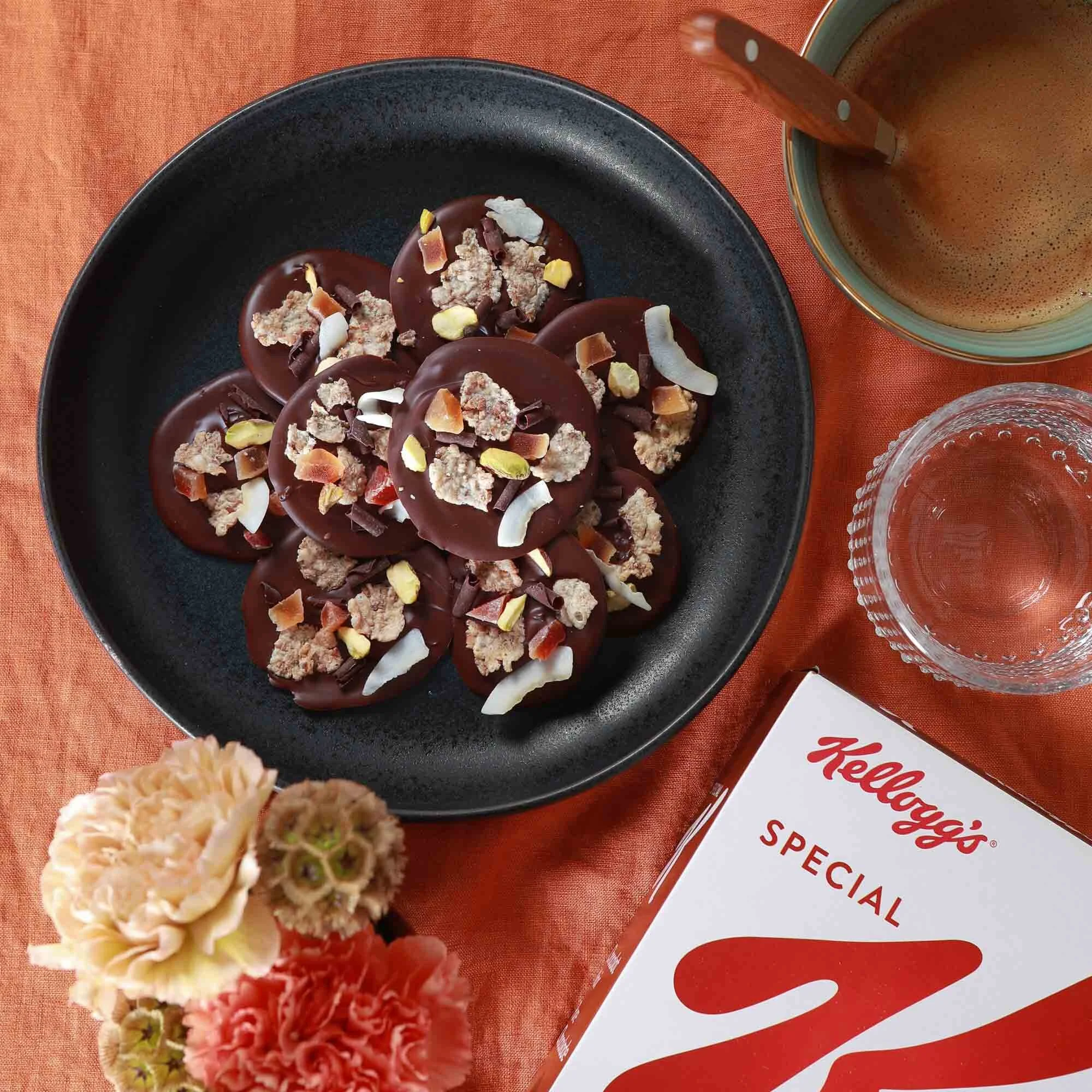 Plaque de chocolat chaud décorée avec des morceaux de chocolat, de la noix de coco, des amandes, et d'autres garnitures, accompagnée d'une tasse de café ou de thé, d'un verre d'eau, et d'une boîte de céréales Kellogg's Special K sur une nappe orange.