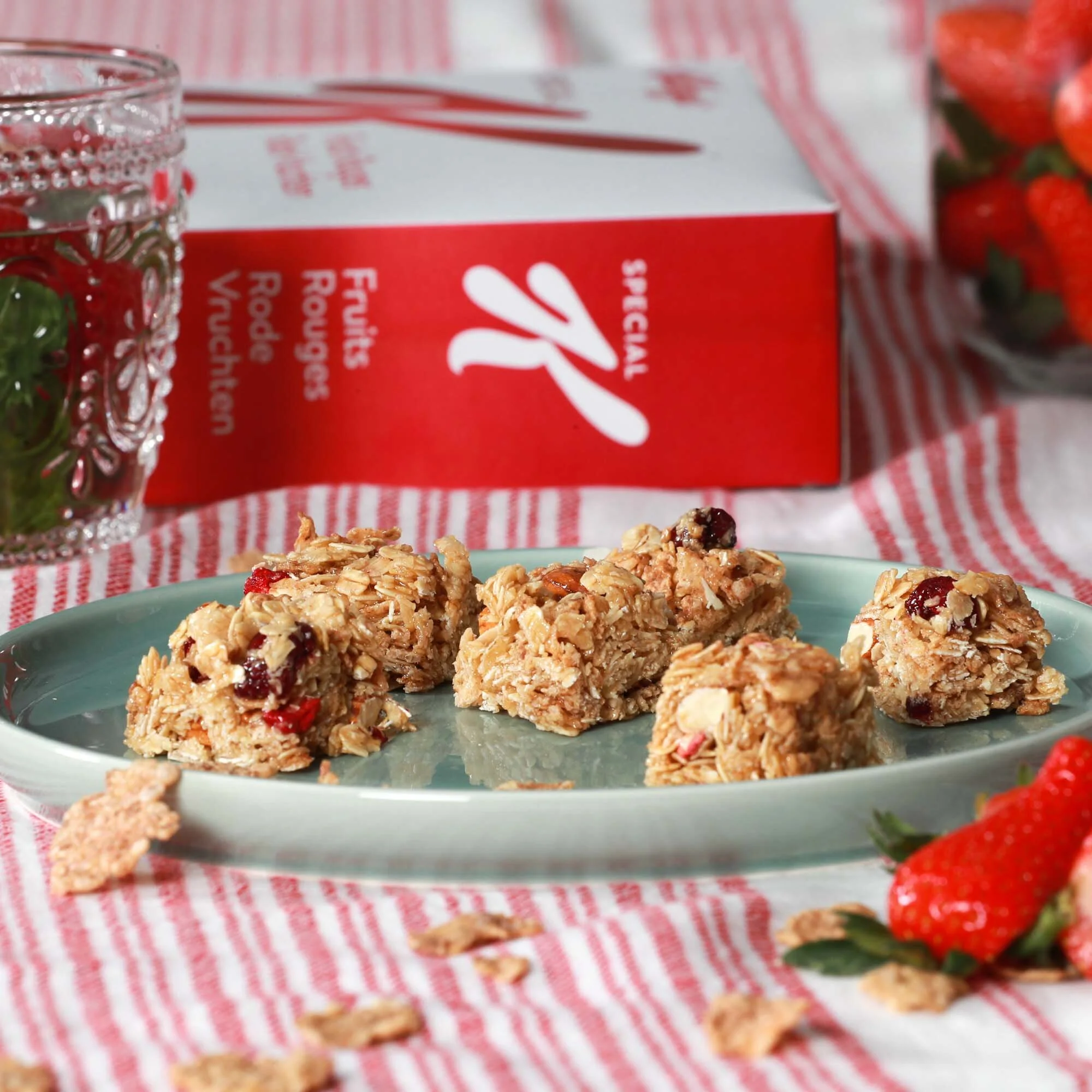 Barres de granola aux fruits rouges sur une assiette, avec une boîte de fruits rouges en arrière-plan et des fraises fraîches à côté, sur une nappe à rayures rouges et blanches.
