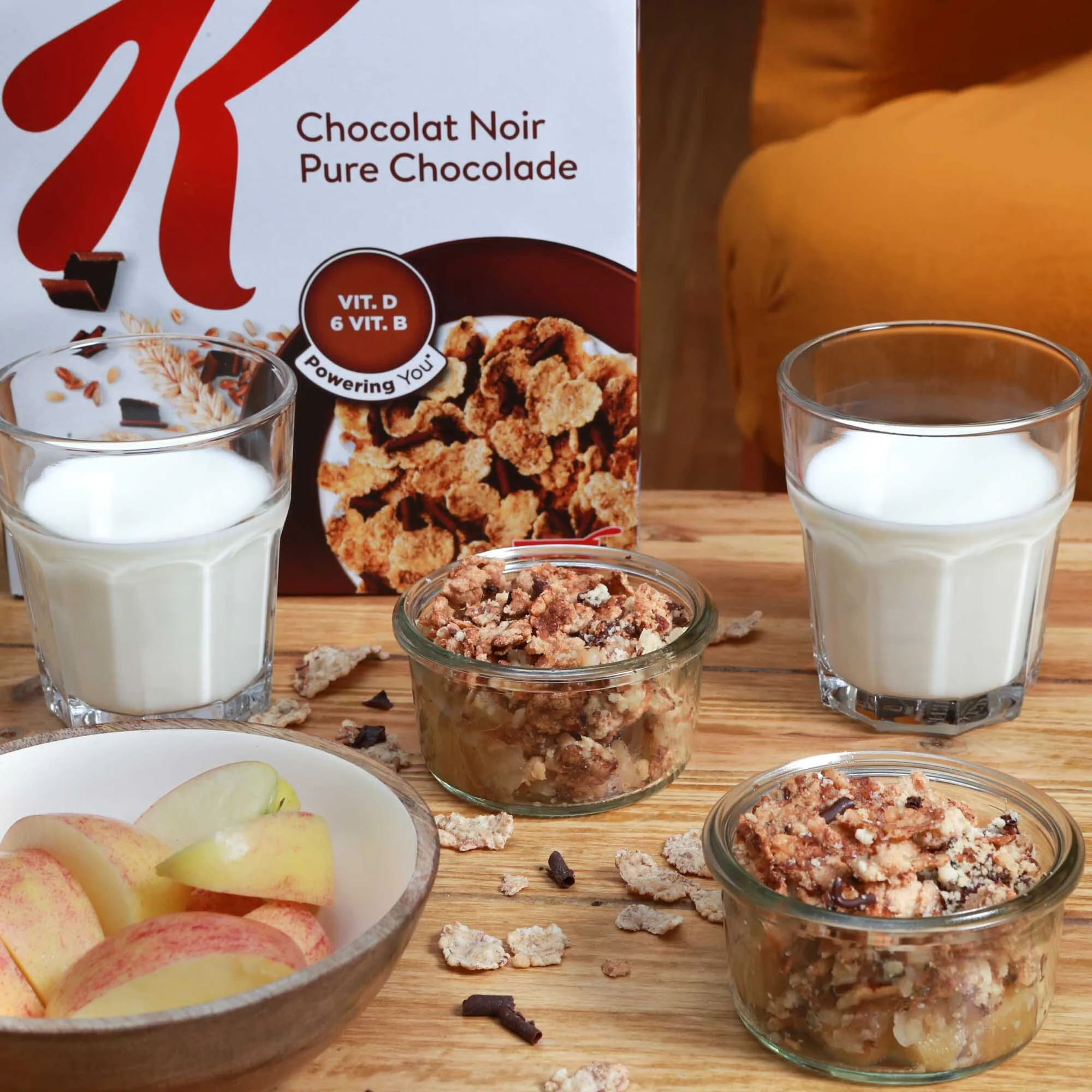 Une boîte de chocolat noir pour petit déjeuner, deux verres de lait, deux pots de crumble aux pommes et chocolat, un bol de tranches de pommes, et quelques miettes et morceaux de crumble sur une table en bois.