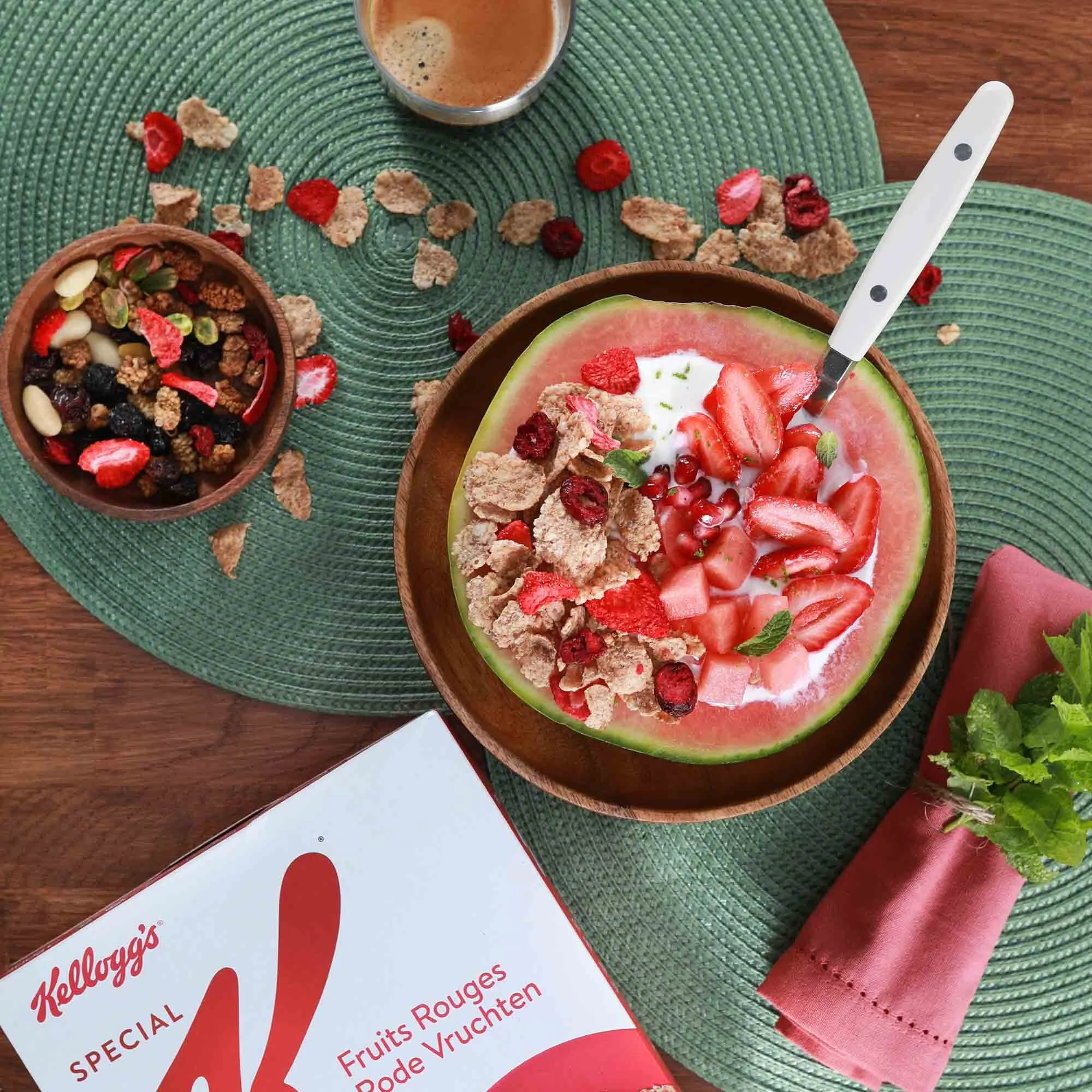 Bol de melon rempli de fraises coupées, comme recette de fruits rouges, avec des céréales et du yaourt. Une tasse de café et un bol de céréales aux fruits rouges également sur une nappe verte. Il y a aussi une boîte de céréales Kellogg's Special aux 