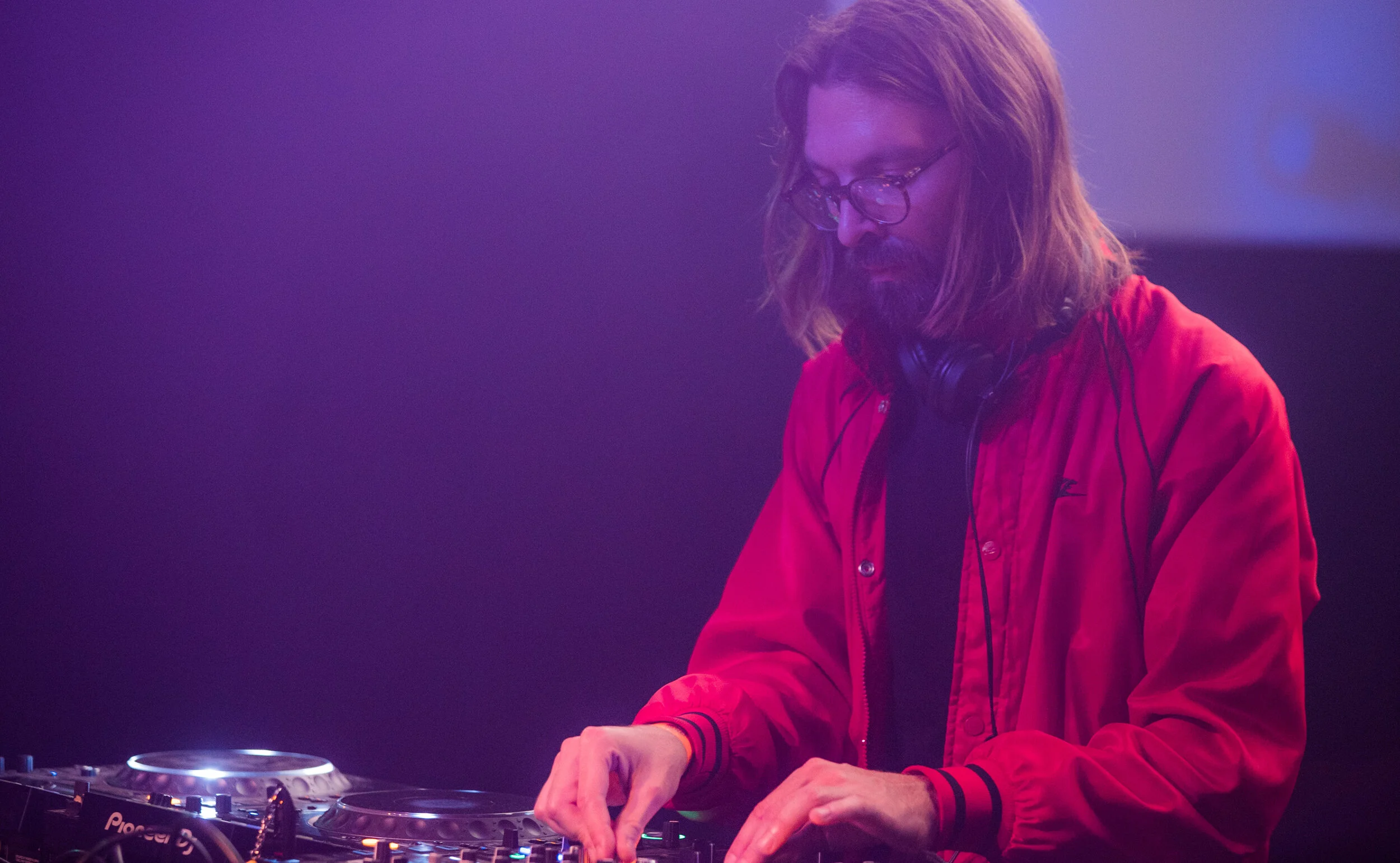 Un DJ avec des cheveux longs, portant des lunettes et une veste rouge, mixant avec un équipement de DJ dans une ambiance sombre avec des lumières violettes.
