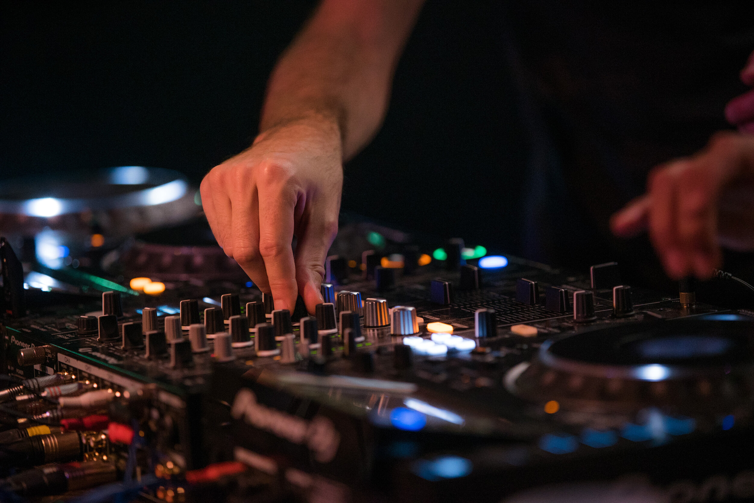 Main d'un DJ manipulant un mélangeur de musique avec des boutons et des cadrans lumineux, dans une ambiance sombre.