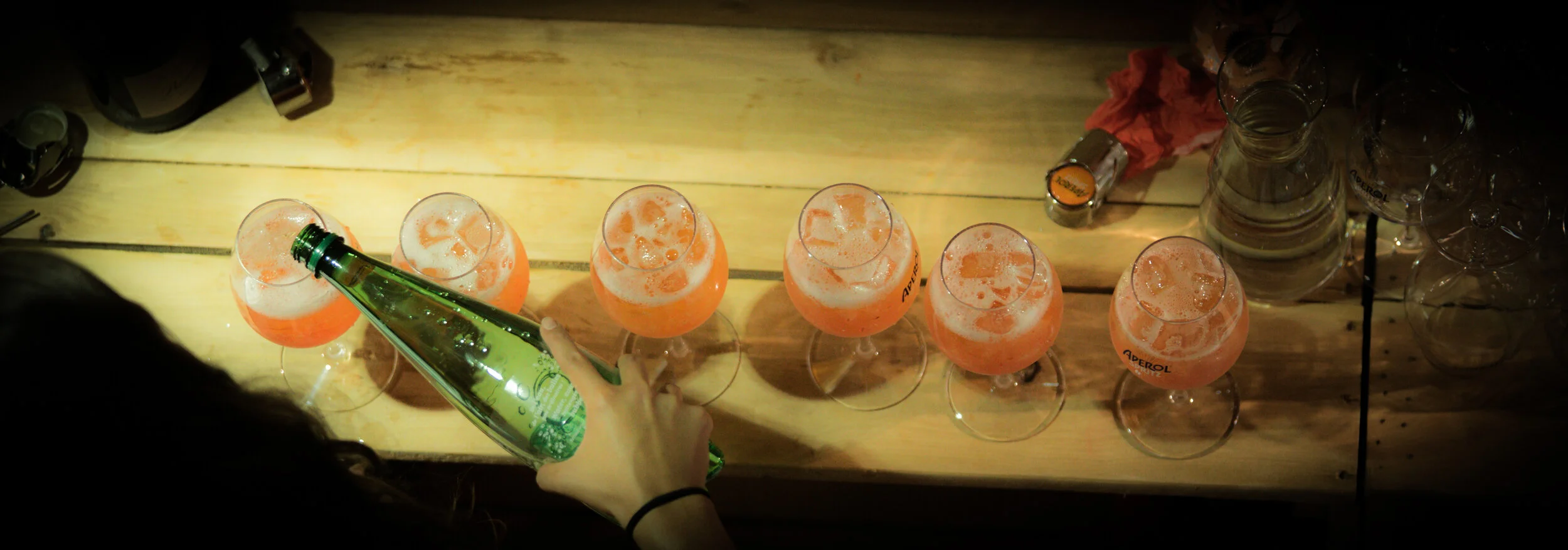 Une personne verse une boisson orange pétillante dans des verres à vin, alignés sur une table en bois, dans un contexte de fête ou de rassemblement.