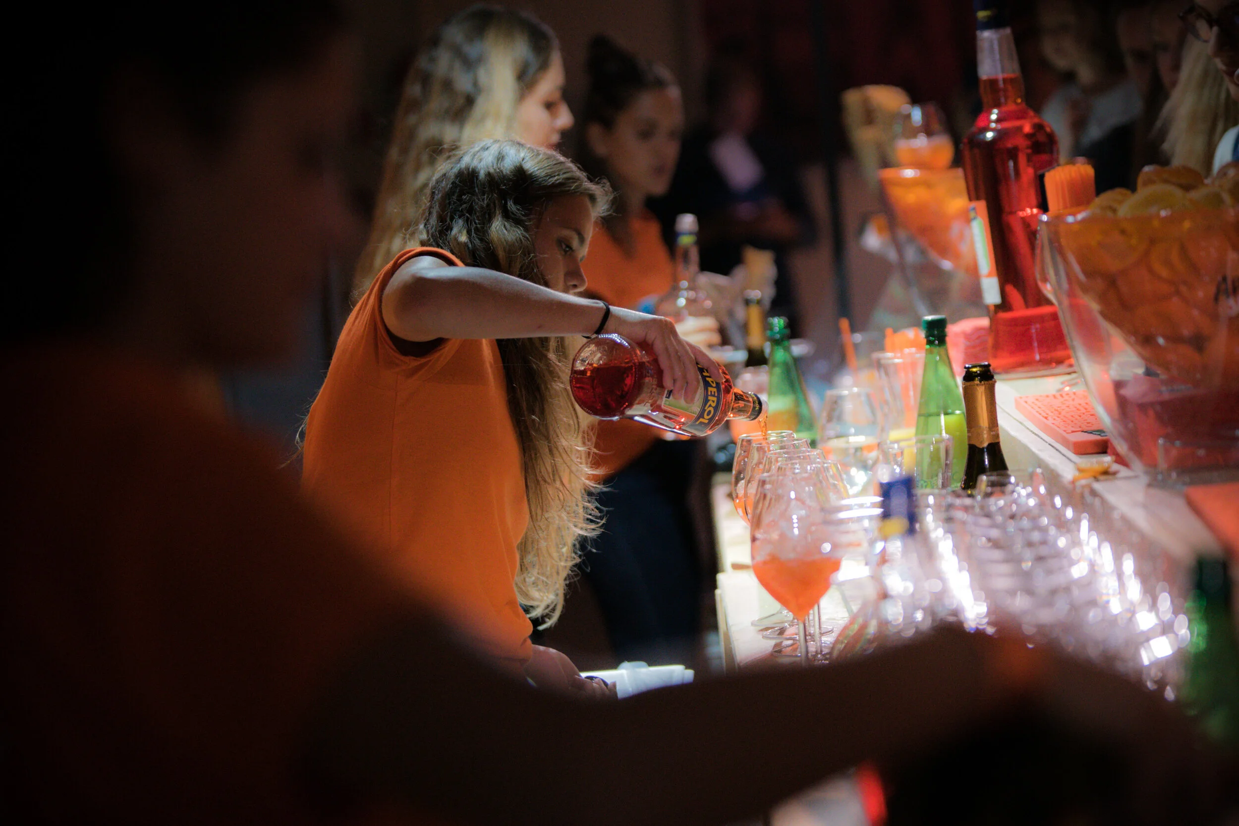 Une jeune femme verse une boisson rouge dans un verre au bar, entourée d'autres personnes et de bouteilles colorées.