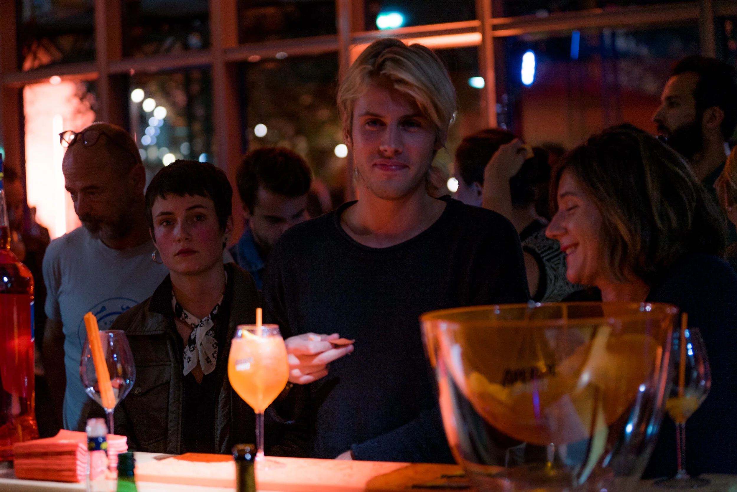Groupe de personnes dans un bar ou un club, ambiance nocturne avec éclairages colorés, une personne blonde au centre tient un téléphone, autres personnes discutant ou ayant des boissons à la main.