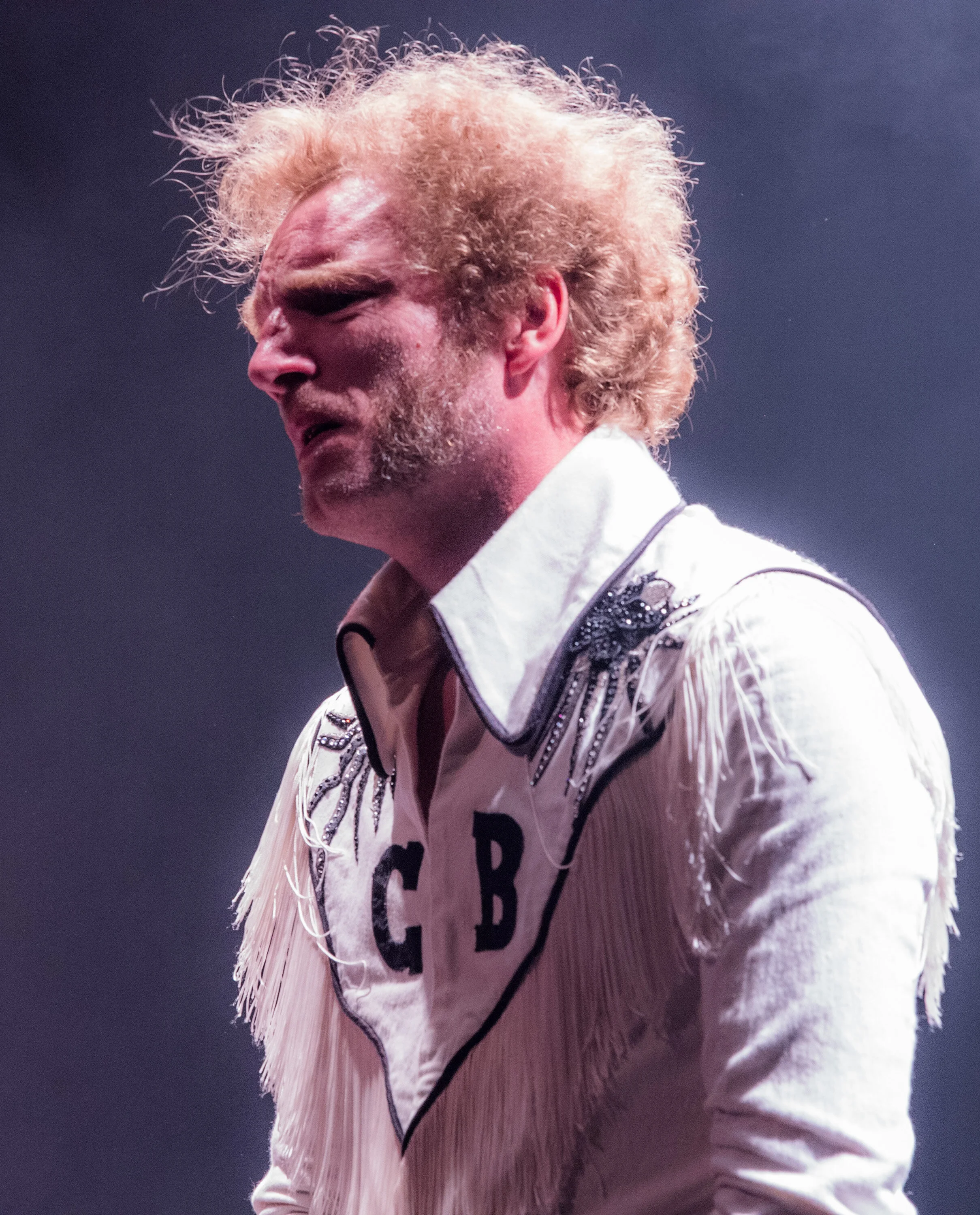 Un homme avec des cheveux bouclés et une barbe, portant une veste blanche avec des détails en galons, lors d'une performance ou spectacle. Le fond est sombre.