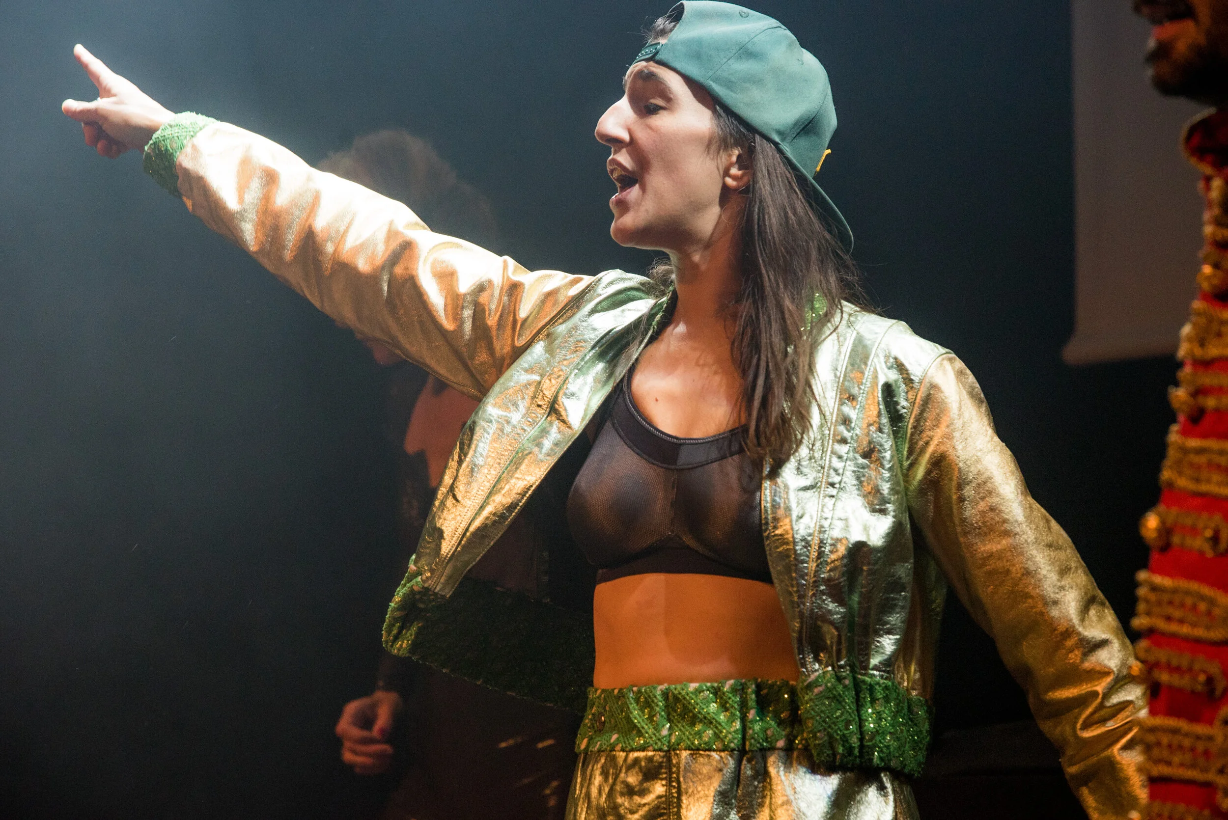 Une femme portant une veste métallique dorée, un crop top noir, un chapeau vert, et des vêtements assortis en fête ou spectacle, pointant du doigt, avec un décor sombre en arrière-plan.