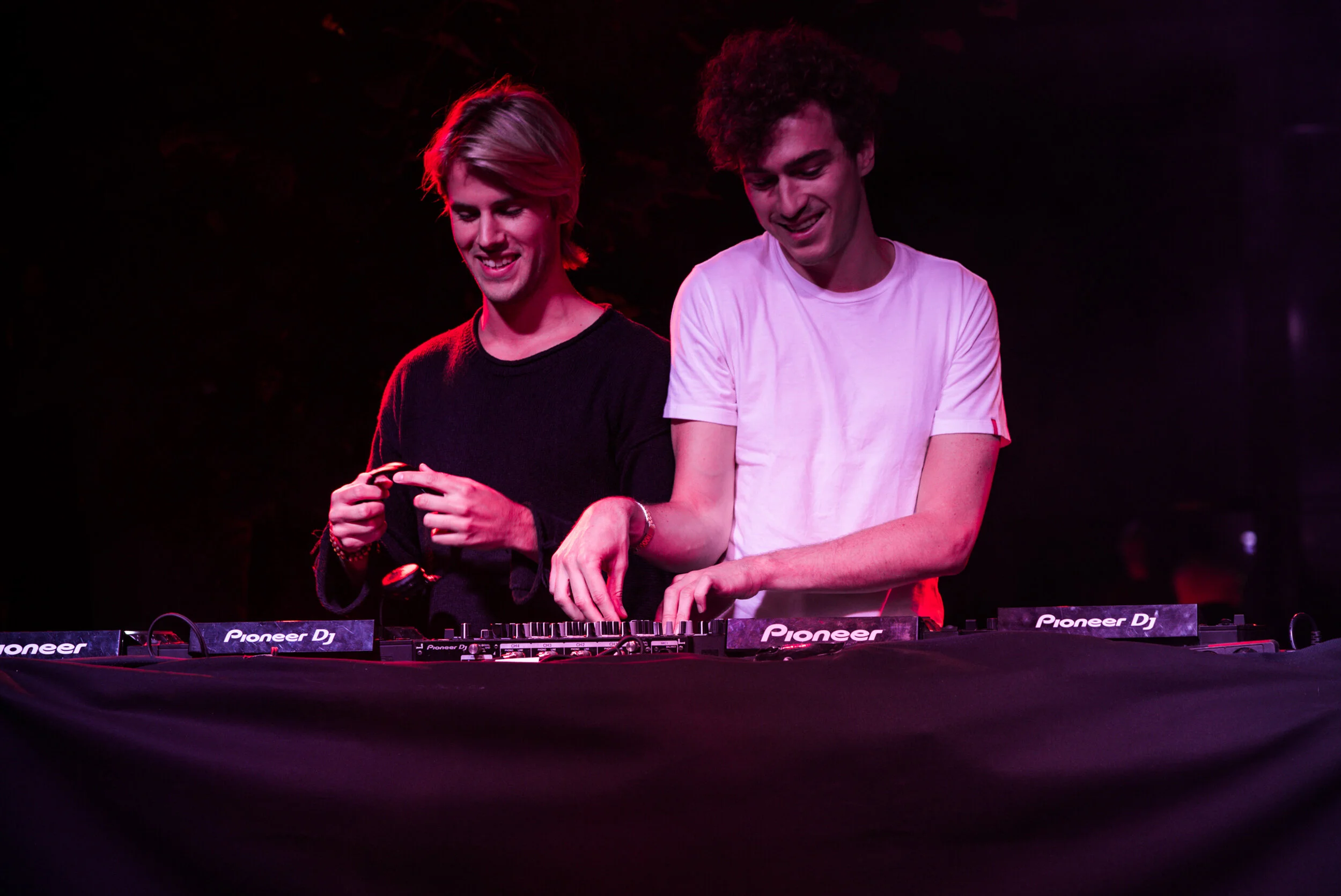 Deux jeunes hommes jouent de la musique sur un DJ Pioneer dans un environnement sombre avec un éclairage violet et rouge.