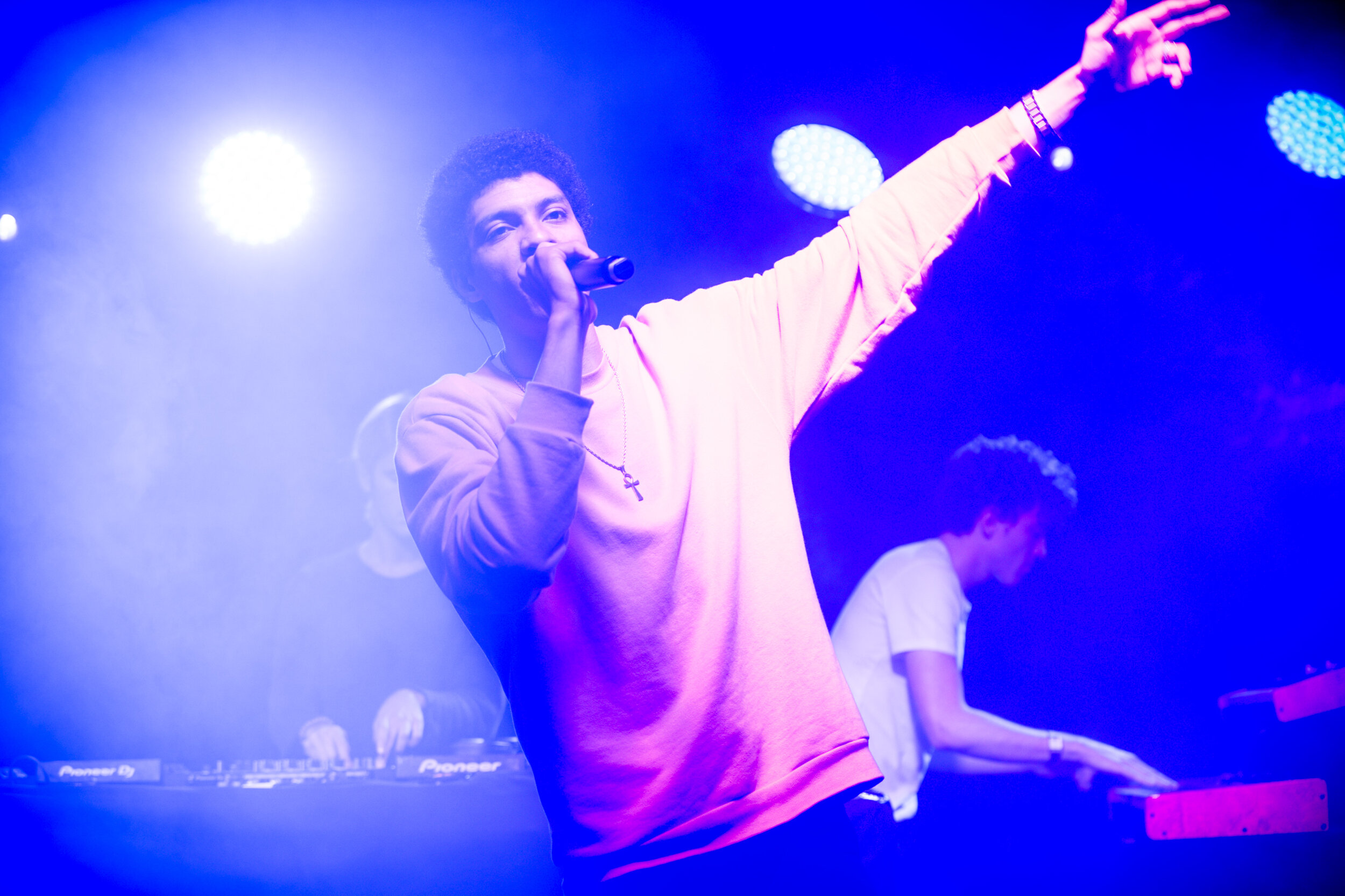 Un chanteur avec un micro, luttant contre un fond de lumières colorées et un DJ, en train de performer sur scène.