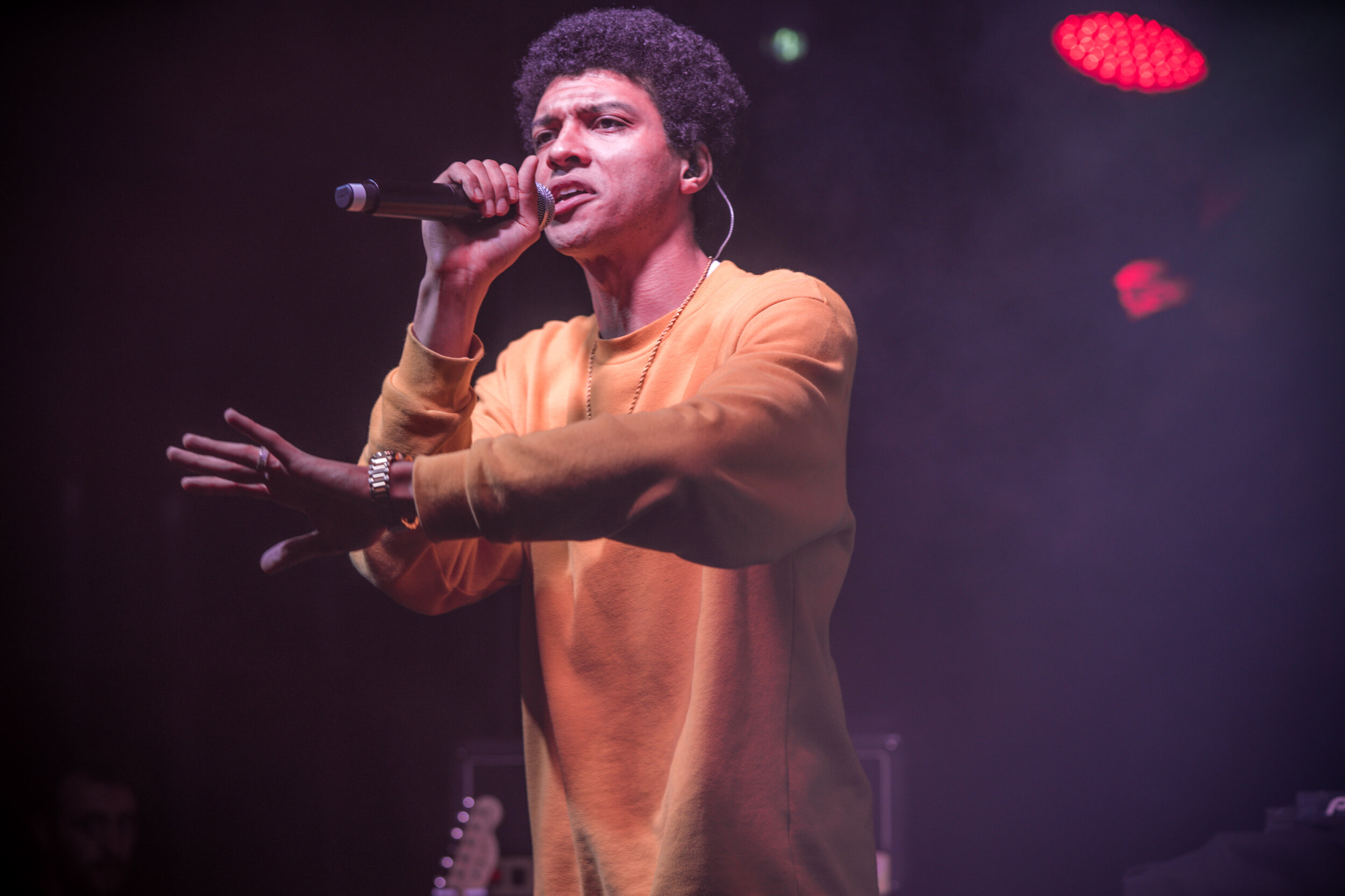 un chanteur noir portant un sweat-orange, tenant un microphone en chantant sur scène avec des lumières rouges en arrière-plan