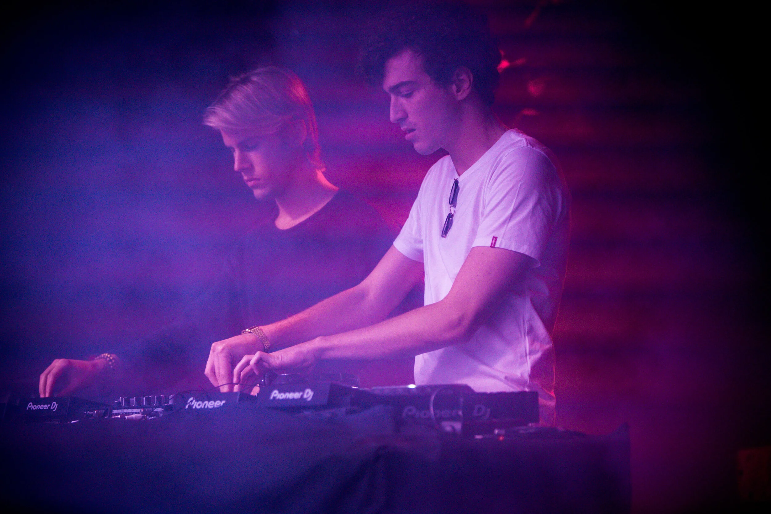 Deux jeunes hommes DJ en train de mixer, éclairés par des lumières violettes et roses, dans une ambiance sombre et brumeuse.