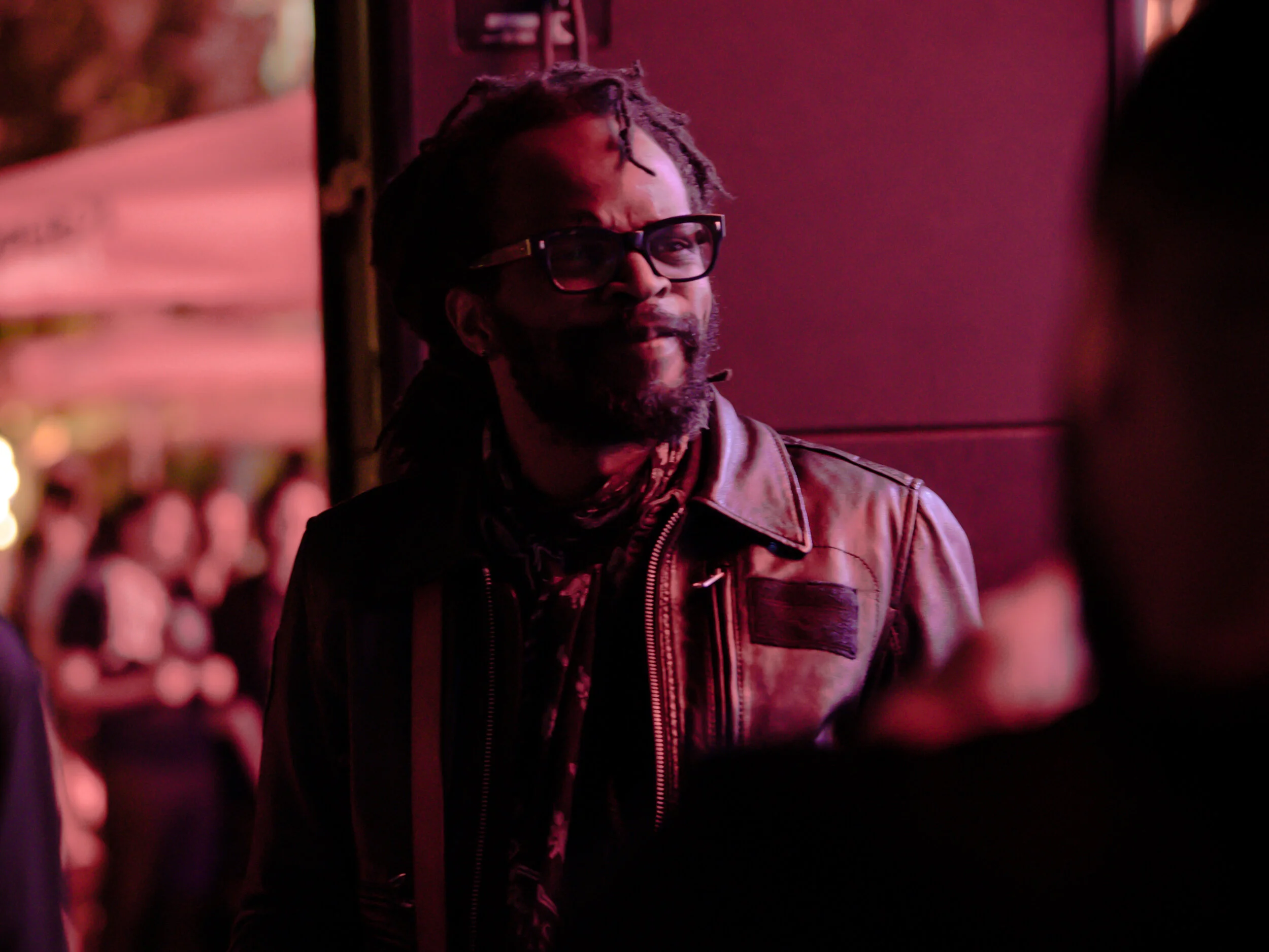Un homme avec des dreadlocks, portant des lunettes, une veste en cuir, et un foulard, dans un environnement lumineux rose ou rouge, entouré d'autres personnes.