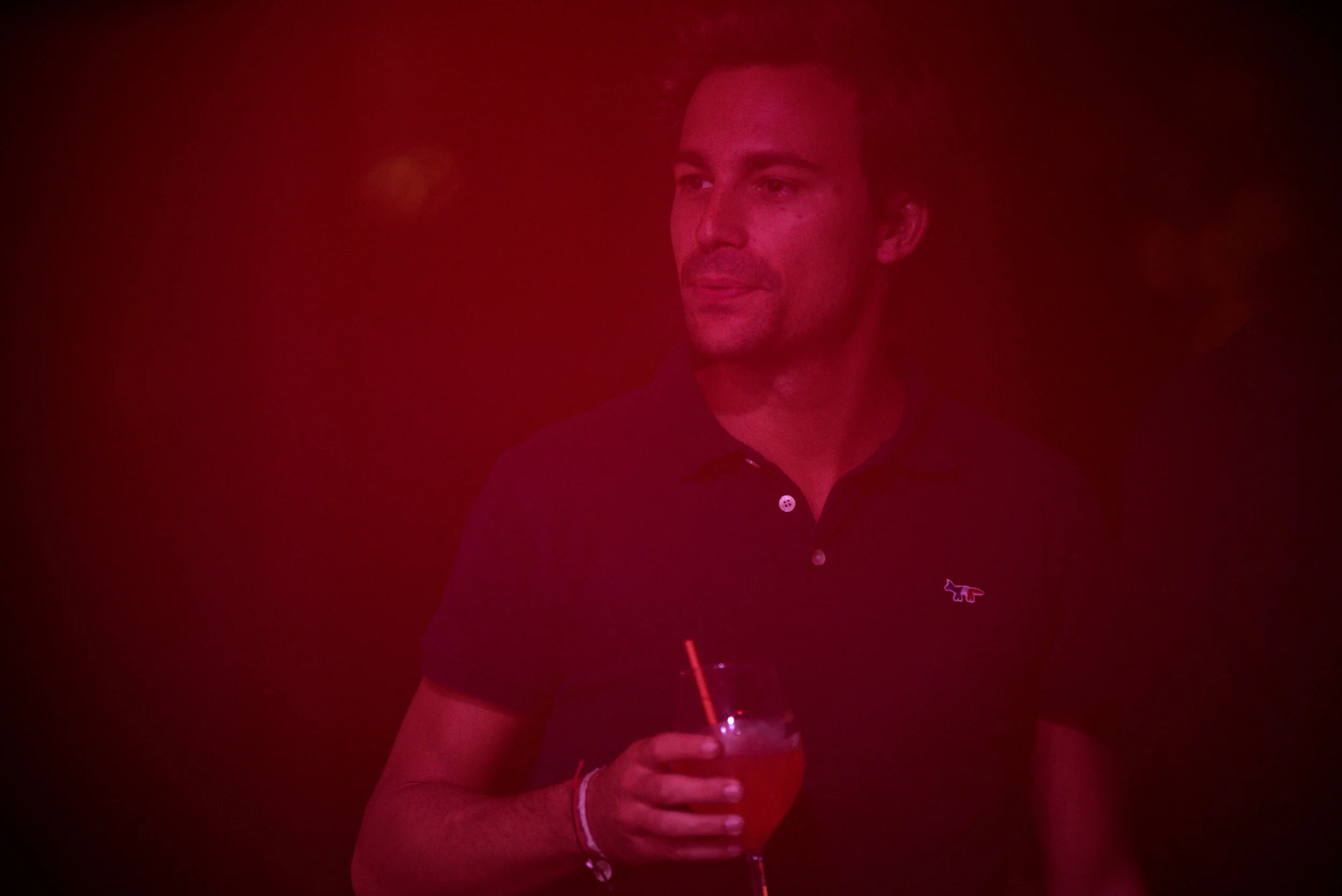 Un homme tenant un verre de cocktail dans une main, portant un polo noir avec un logo de lézard, dans un environnement sombre avec un éclairage rouge.