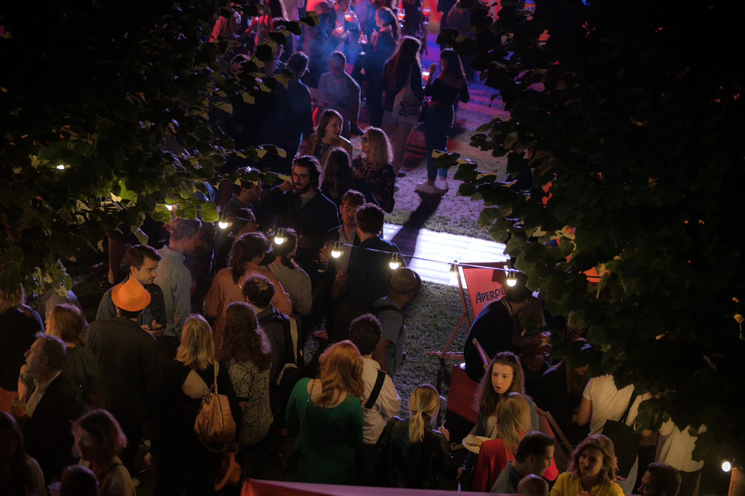 Une foule de personnes assises et debout lors d'une soirée en plein air, éclairée par des lampes suspendues, avec des arbres entourant la scène.