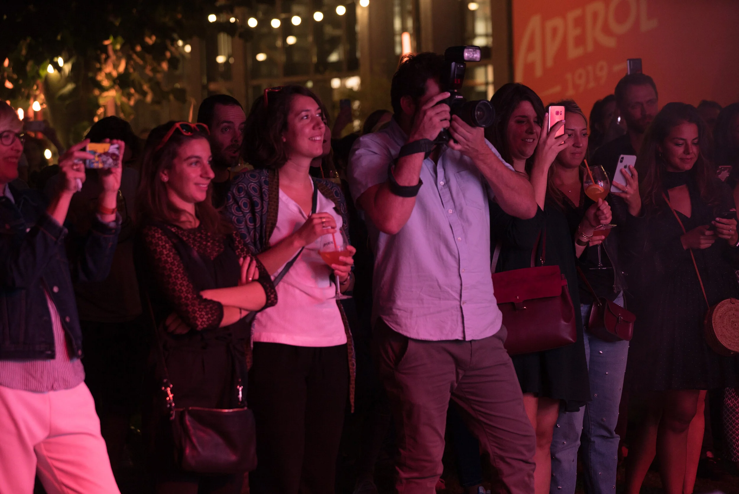 Groupe de personnes regardant un spectacle ou un événement, certains prennent des photos ou vidéos, ambiance festive avec éclairage rose.
