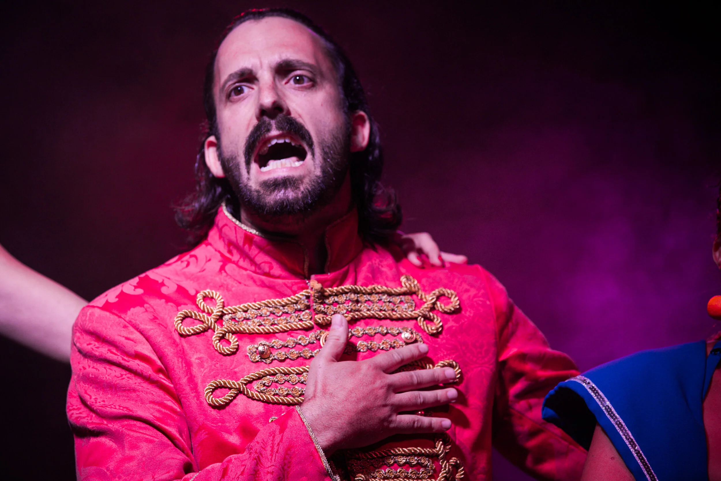 Un homme avec des cheveux longs et une barbe, portant un costume rouge avec des détails dorés, chante ou parle avec intensité, main sur la poitrine, dans un contexte de scène ou de performance.