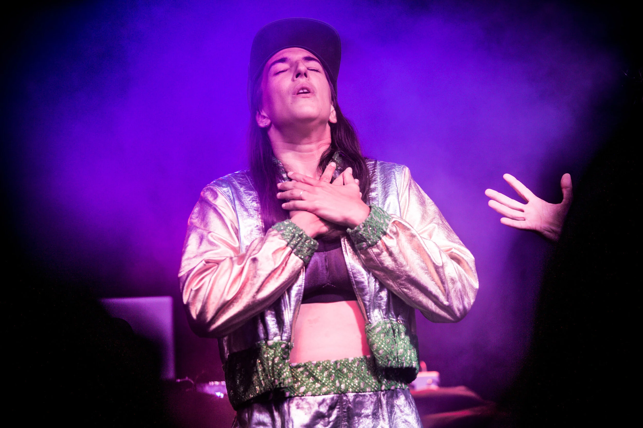 Une jeune femme habillée en costume argenté avec des détails verts, portant une casquette noire, exprime une émotion intense en se tenant la main sur la poitrine lors d'une performance scénique, avec un fond violet et une main levée à droite.