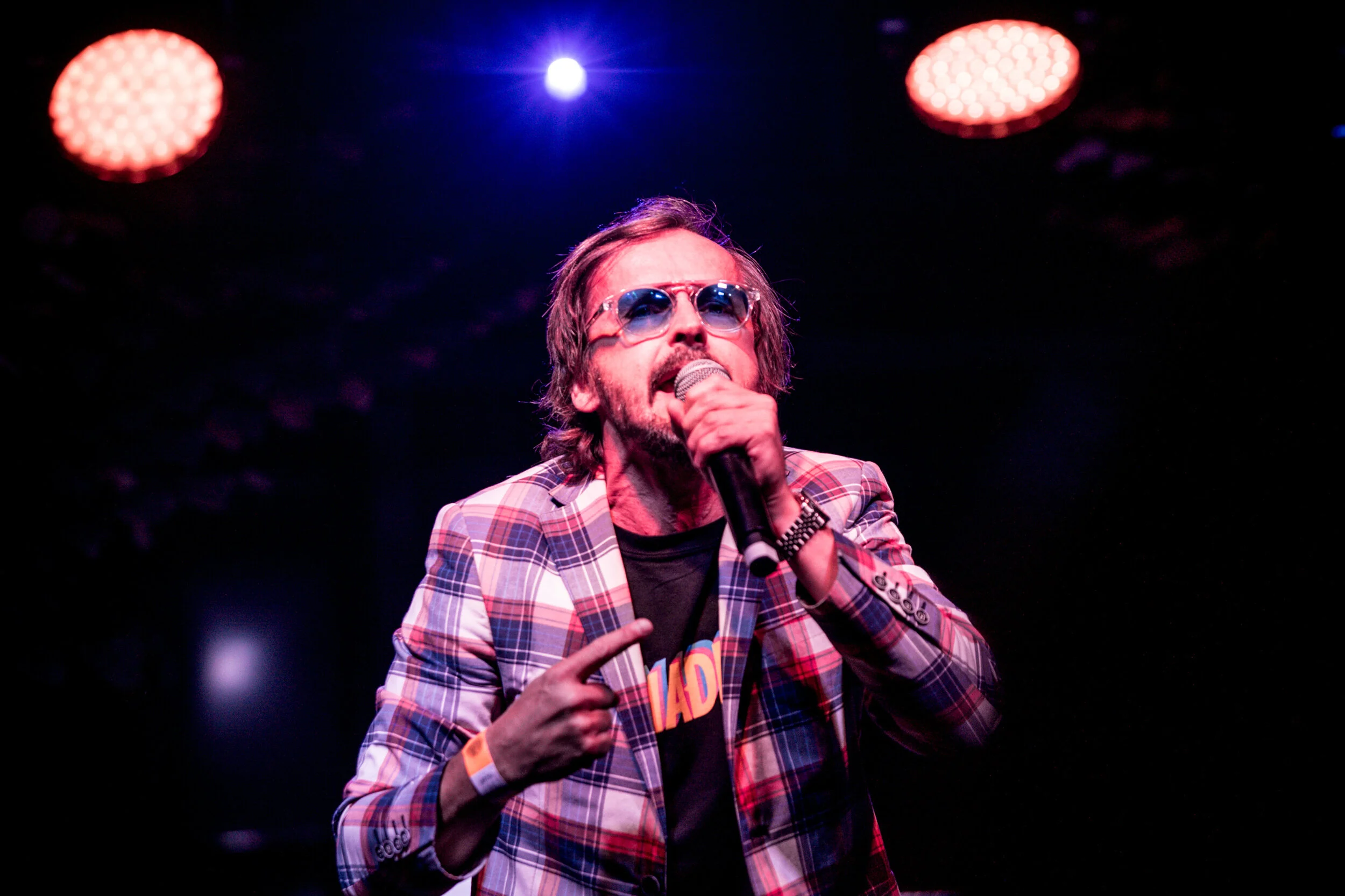 Un chanteur avec des lunettes de soleil, un blazer à carreaux et un t-shirt noir, chantant ou parlant dans un microphone lors d'une performance sur scène sous des lumières colorées.