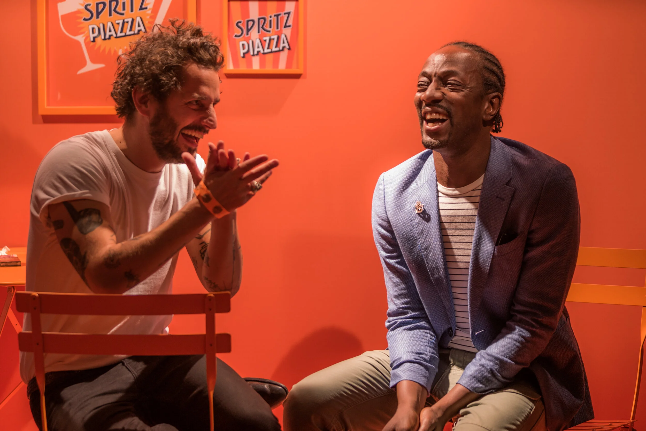 Deux hommes rient et discutent dans une pièce orange avec des affiches 'Spritz Piazza' au mur.