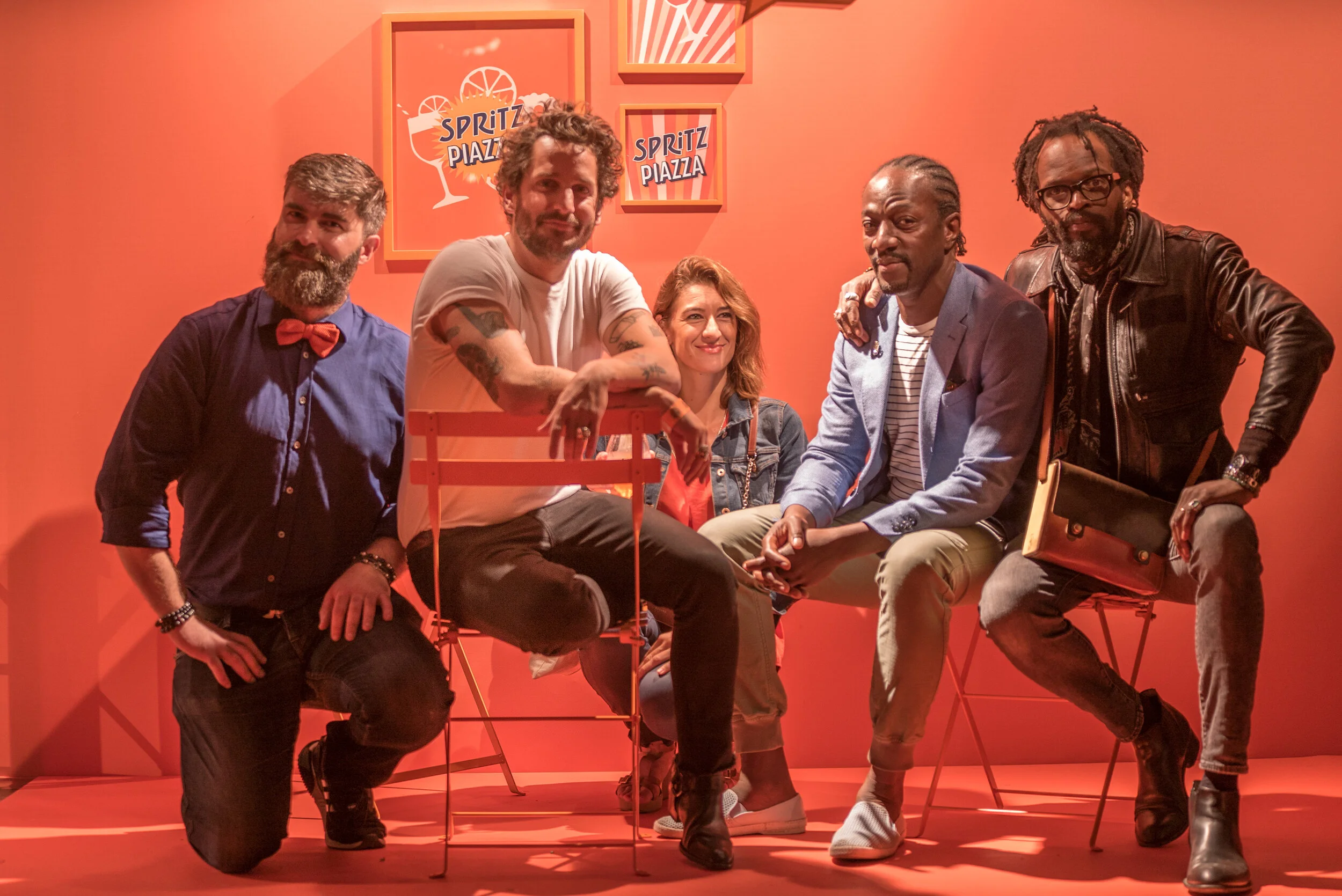 Un groupe de six personnes posant dans un décor rose avec des panneaux 'Spritz Piazza'.