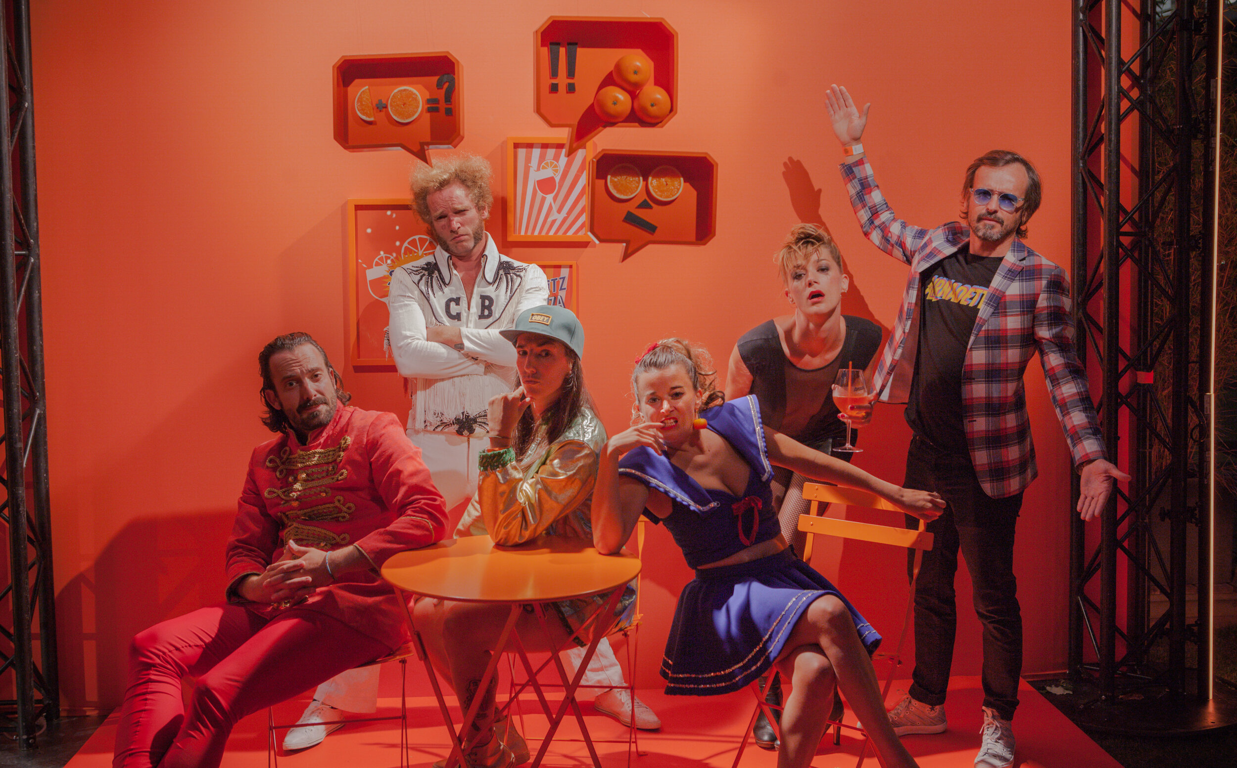 Groupe de sept personnes posant devant un mur orange avec des illustrations de fruits dans des cadres colorés, certains assis autour d'une table orange, d'autres debout ou assis sur des chaises, portant des tenues variées, dans une scène avec une str