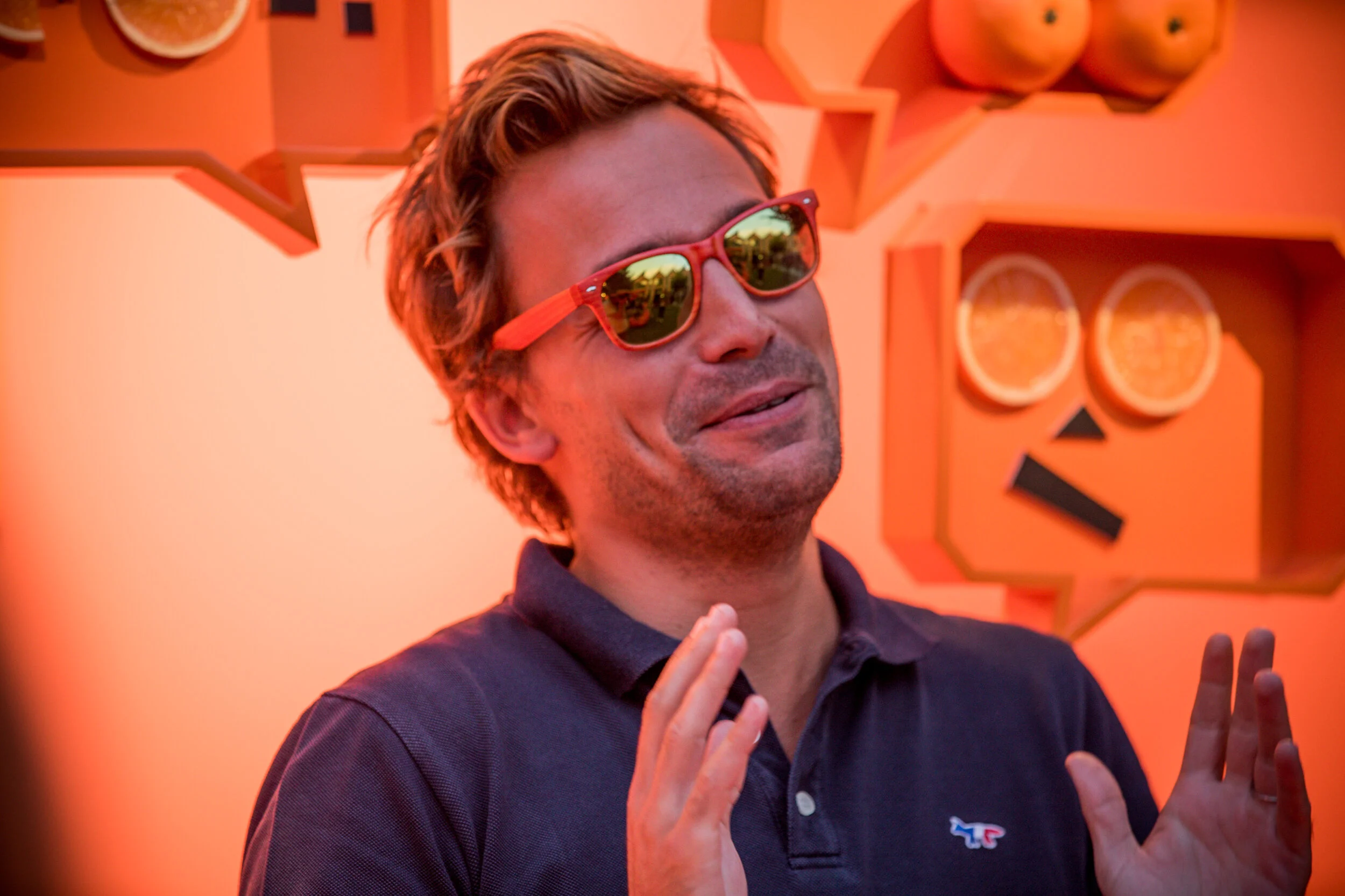 Un homme porte des lunettes de soleil rouges et un polo noir, il sourit et fait un geste avec ses mains. Il est devant un décor orange avec des motifs de fruits et formes géométriques.