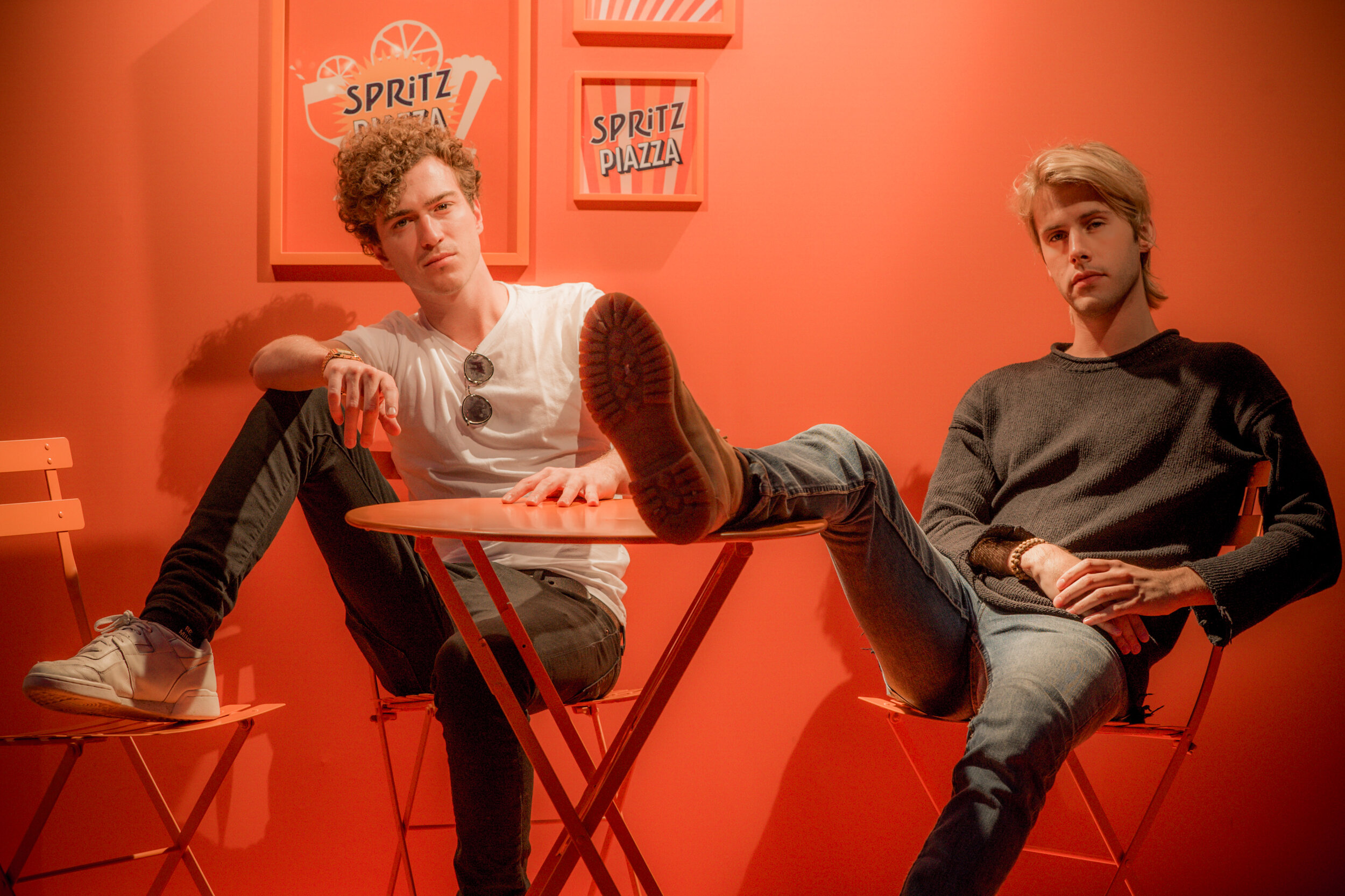 Deux jeunes hommes assis sur des chaises autour d'une petite table dans un décor orange. Ils ont une attitude décontractée. Le mur derrière eux est décoré de posters avec l'inscription « Spritz Piazza ».
