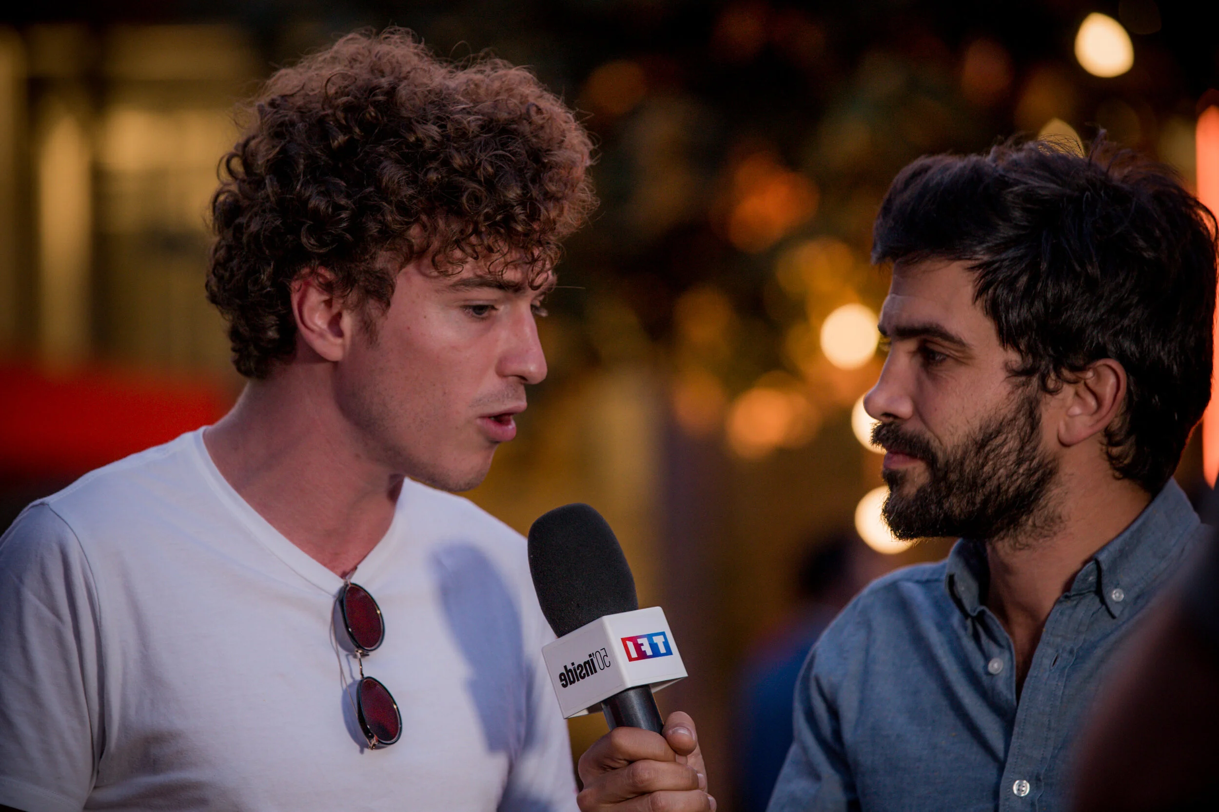 Deux hommes en train d'interviewer, l'un tient un microphone avec le logo TET, dans un environnement nocturne avec des lumières floues en arrière-plan.