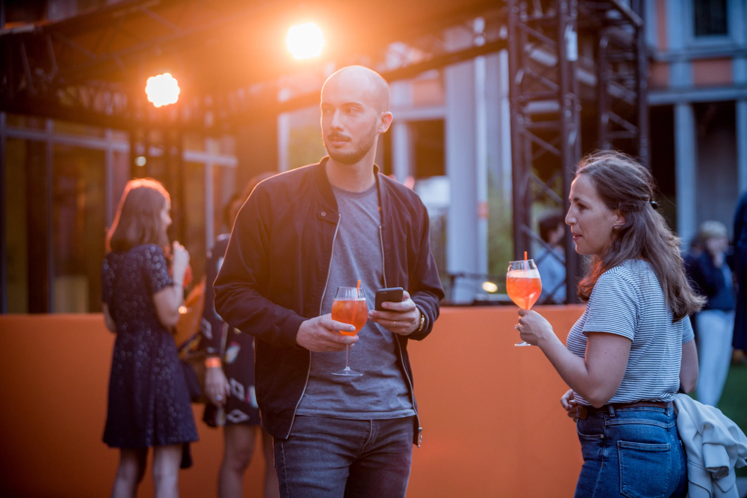 Deux personnes, un homme et une femme, discutent tout en tenant des verres avec des boissons colorées dans un environnement extérieur lors d'une soirée.
