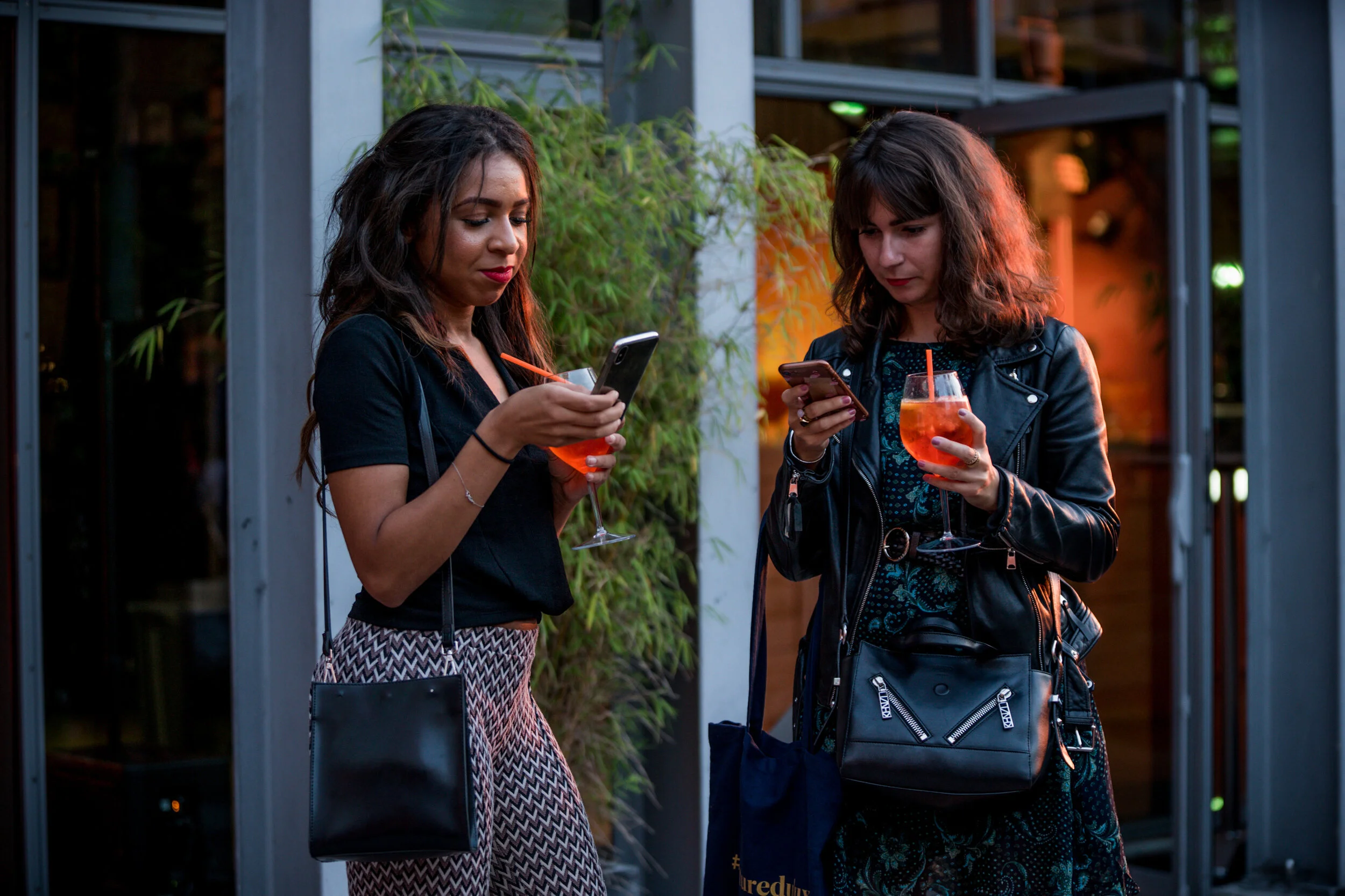 Deux femmes regardant leurs téléphones portables tout en tenant des cocktails rouges avec des pailles, debout près d'une entrée d'un bâtiment moderne avec des plantes.