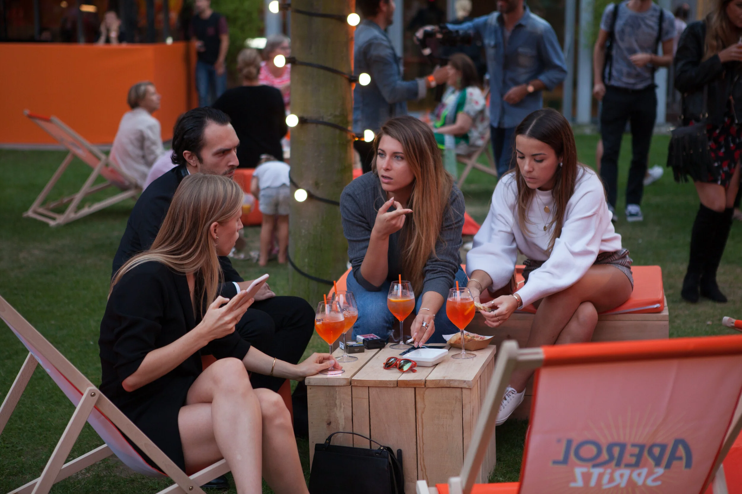 Groupe de quatre jeunes femmes assises autour d'une table basse en bois dans un jardin lors d'une soirée, avec des boissons orange dans des verres, en train de discuter.