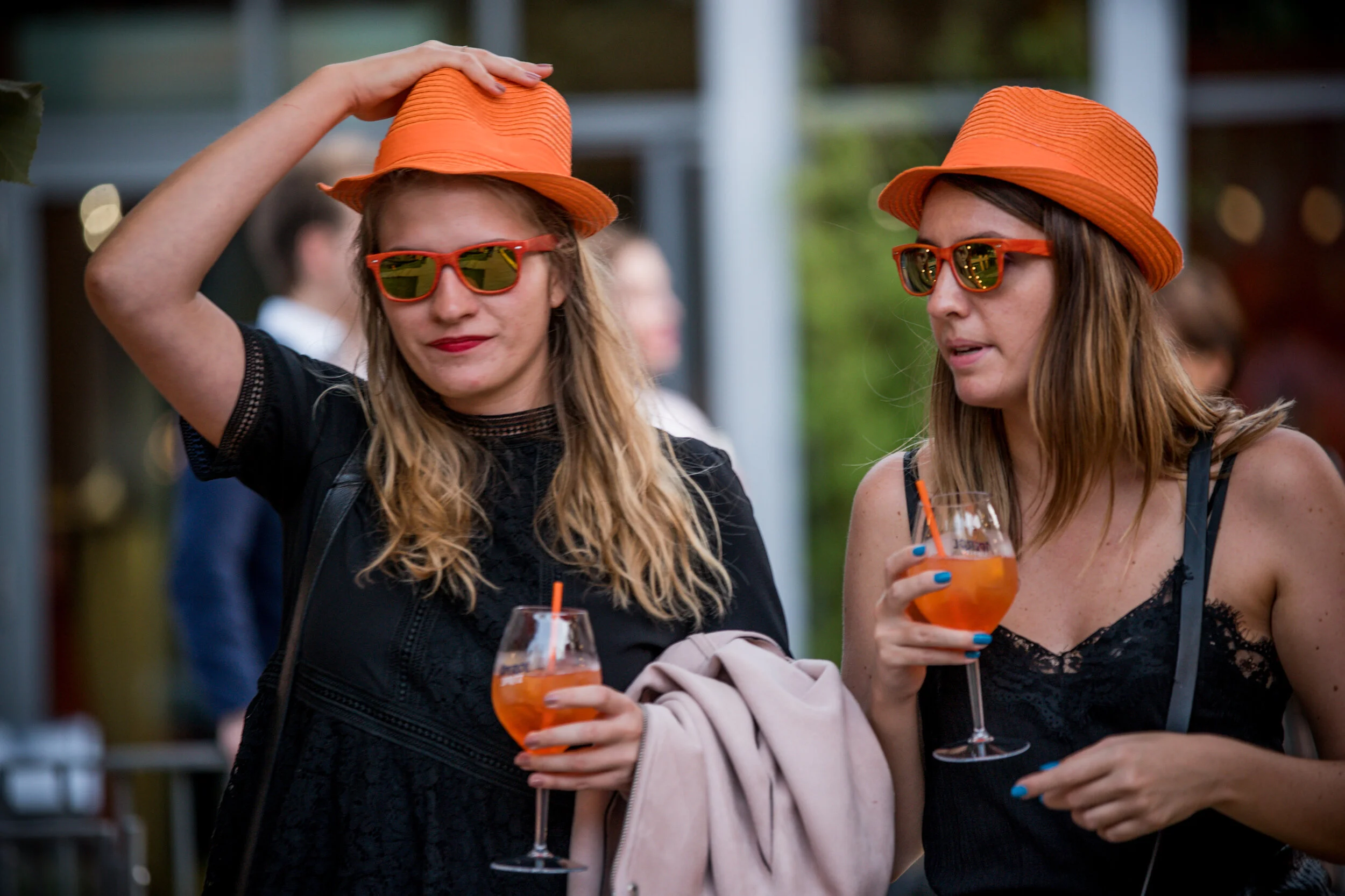 Deux femmes portent des chapeaux orange et des lunettes de soleil orange. Elles tiennent des verres de cocktail orange et semblent parler lors d'une fête en plein air.