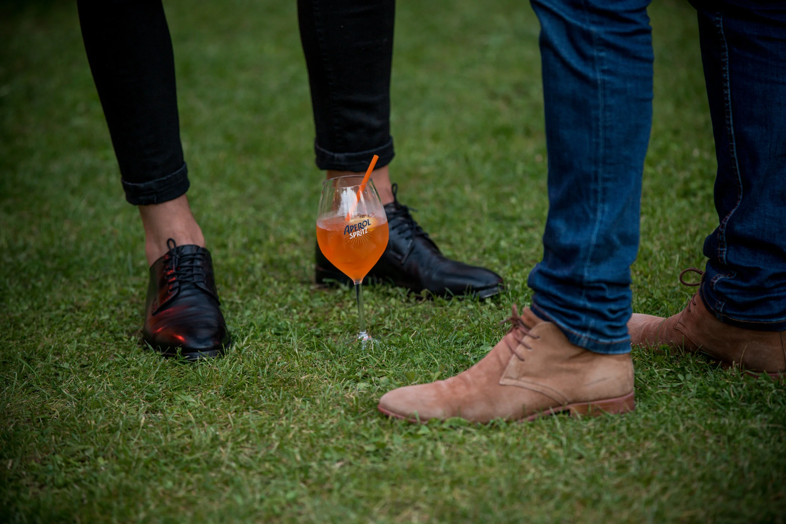 Gens portant des chaussures différentes, avec un verre d'Apérol Spritz posé sur le sol entre eux, sur de l'herbe