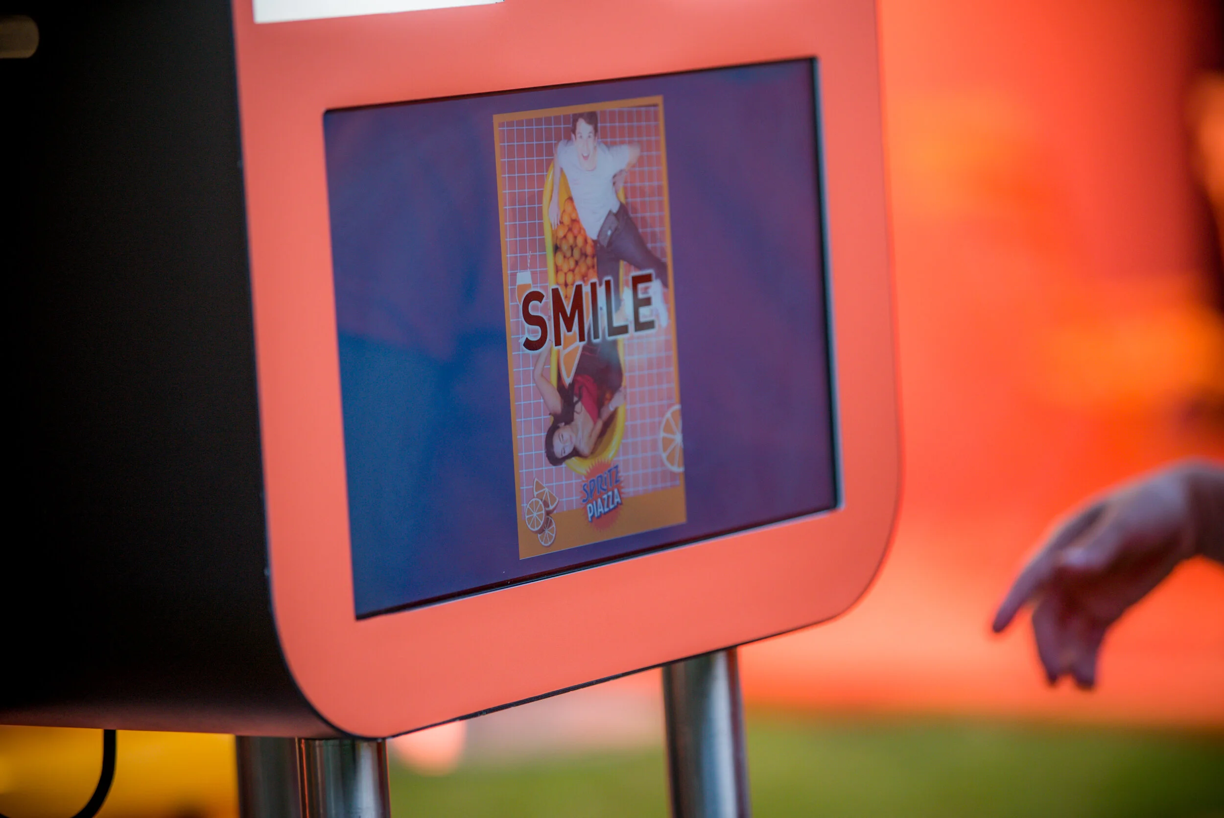 Une borne interactive orange avec un écran affichant une publicité colorée pour une boisson gazeuse avec le mot "SMILE" et des images de jeunes gens s'amusant.