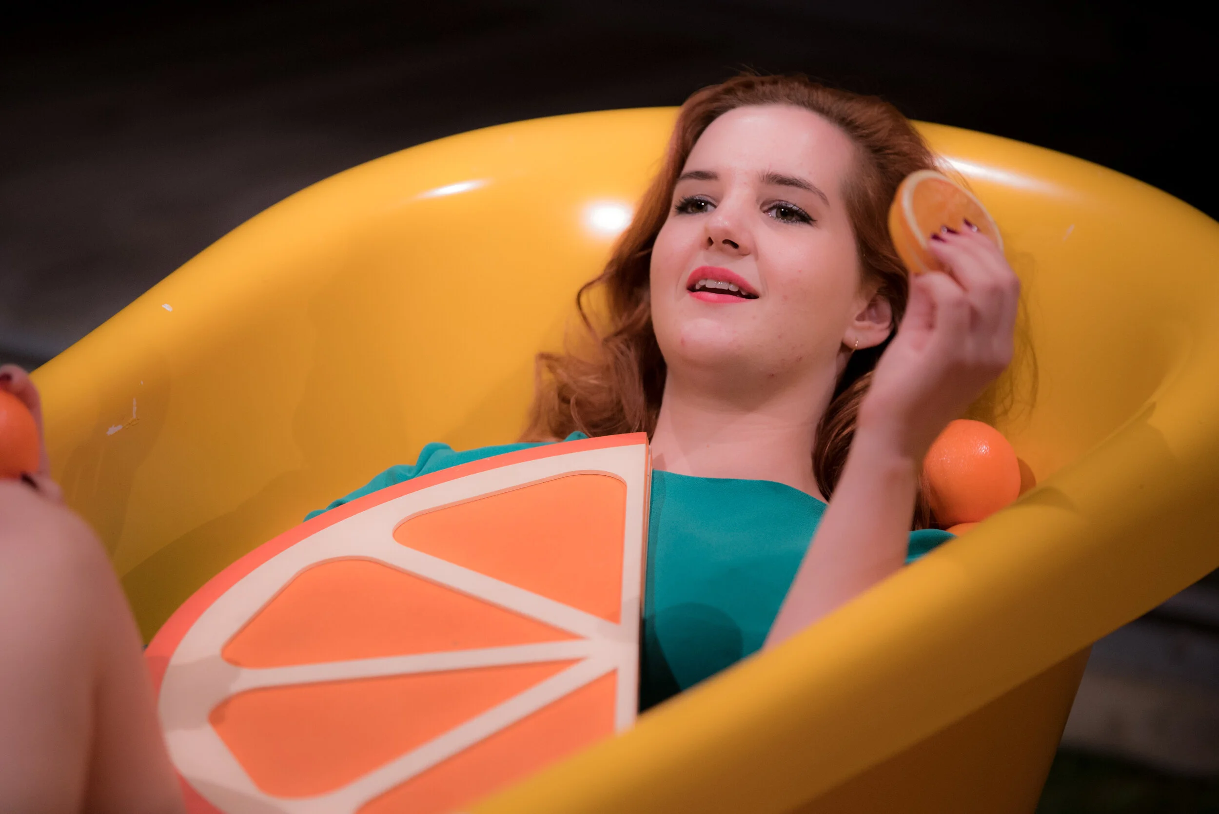 Une jeune femme avec des cheveux ondulés rouges allongée dans une baignoire gonflable jaune, tenant une tranche d'orange et un demi-orange sur sa tête, avec une autre orange à ses côtés.