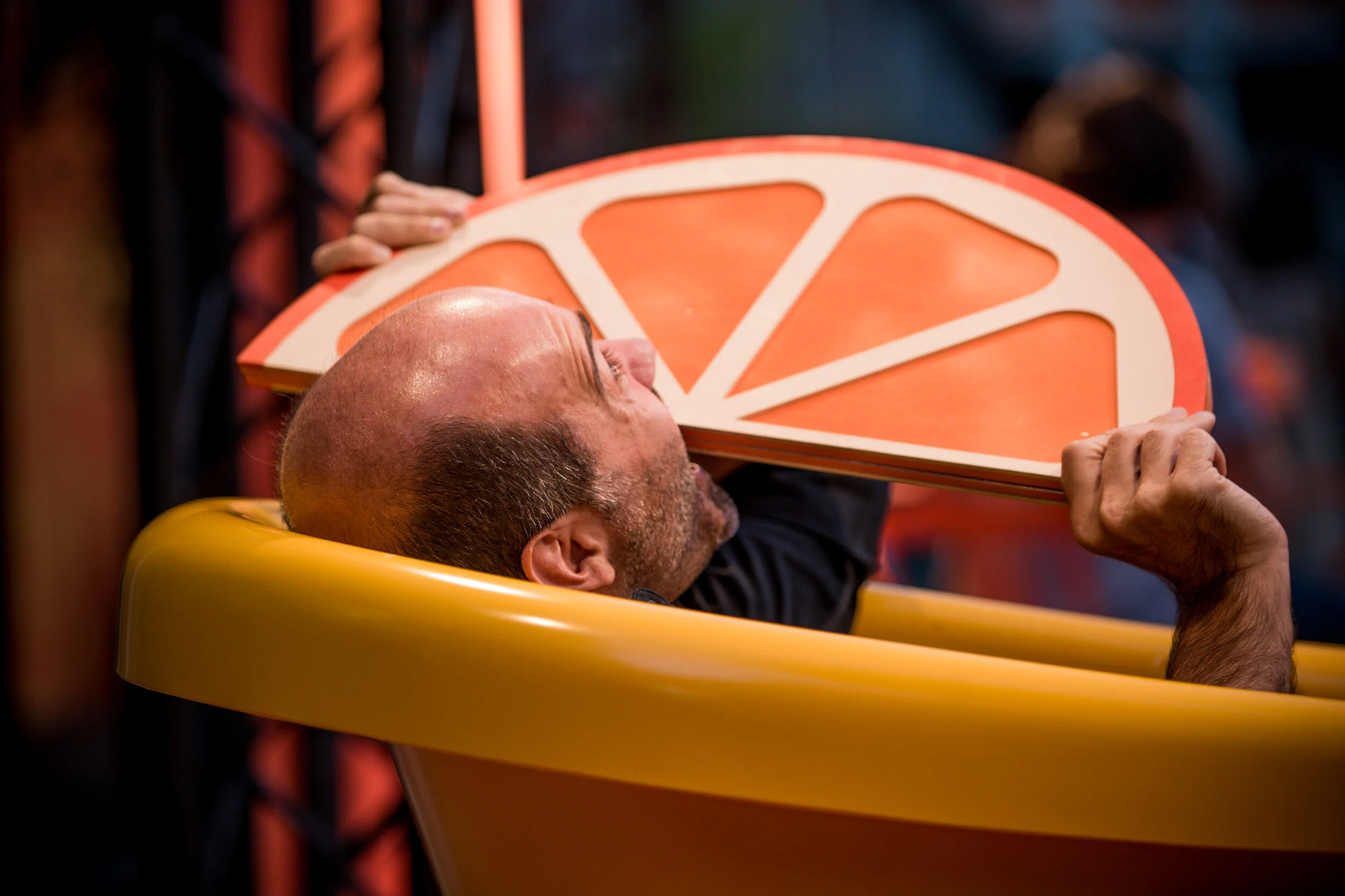 Un homme dans une chaise haute, tenant une pièce en forme de tranche de citron, appuyée sur sa bouche