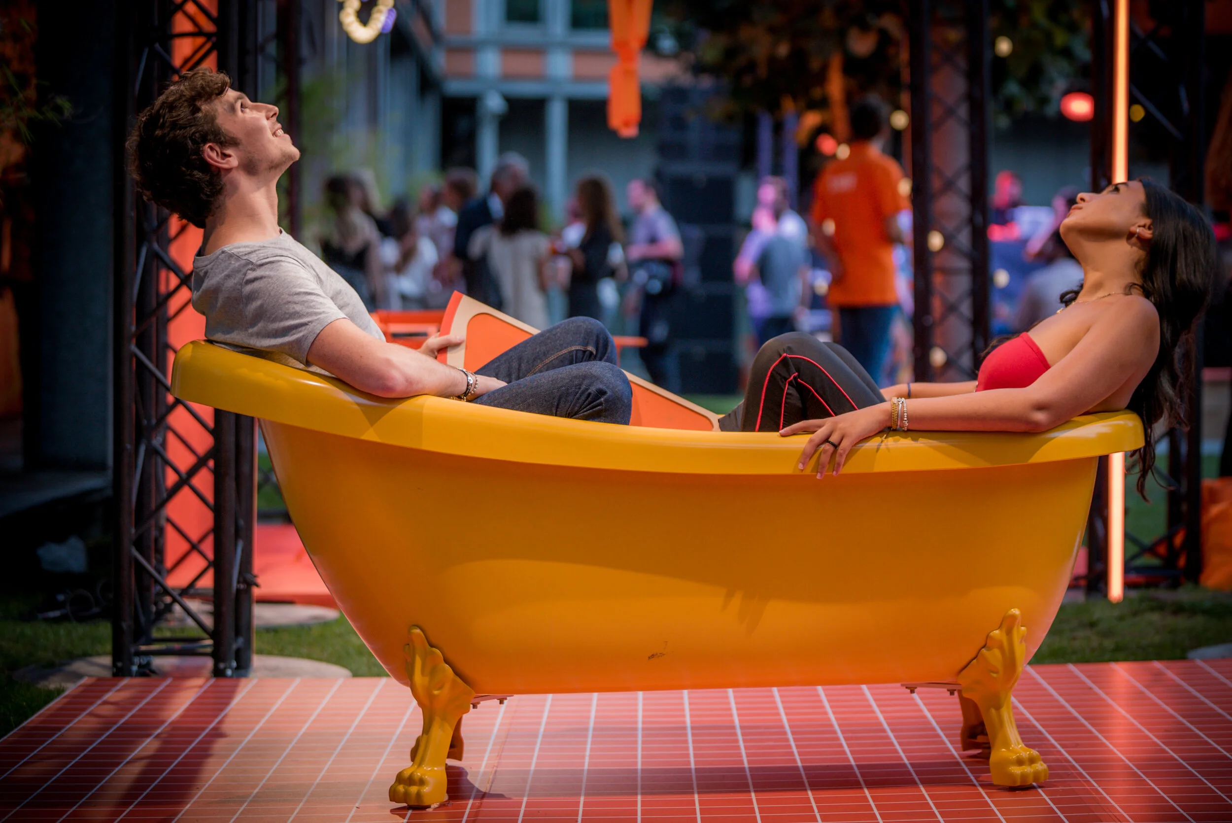 Deux personnes sont allongées dans une baignoire jaune, souriant et regardant vers le haut. Elles sont dans un environnement lumineux avec des personnes en arrière-plan lors d'un événement social ou un festival.