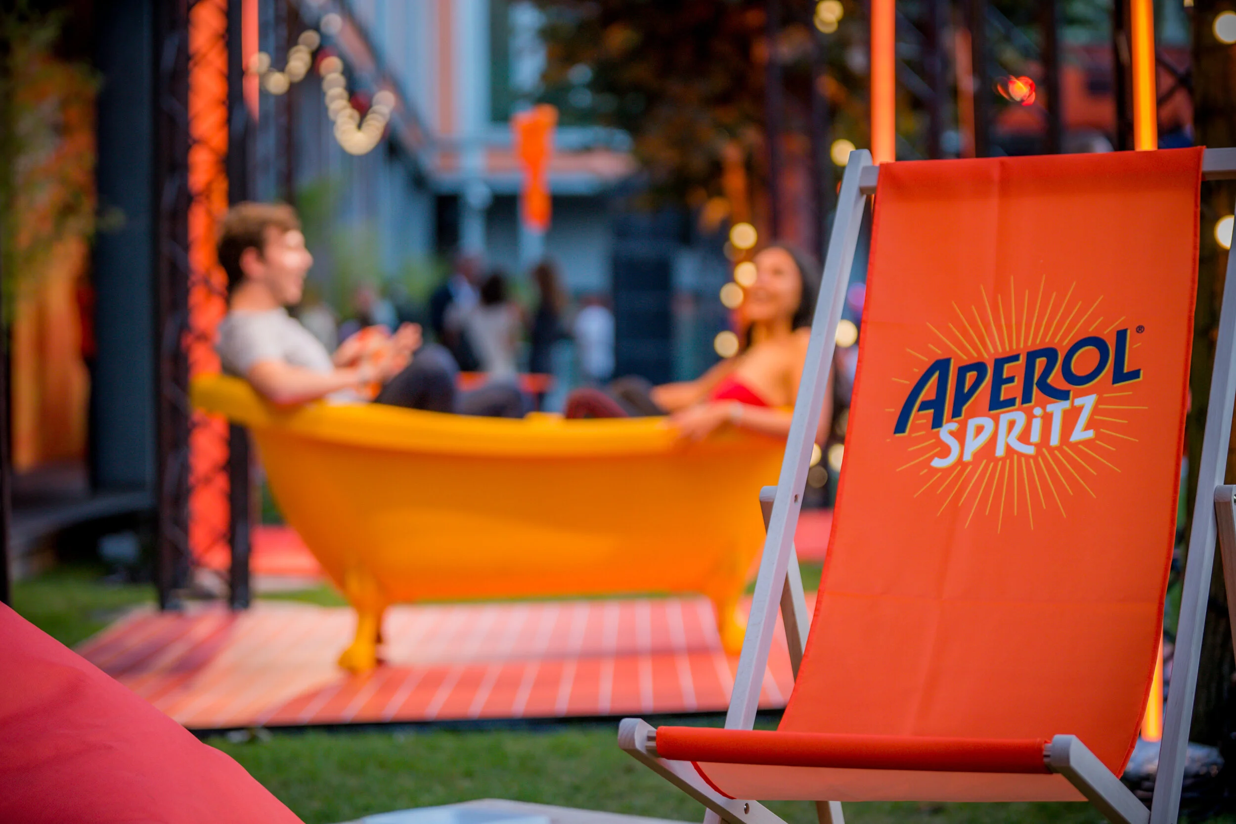 Une chaise longue orange avec le logo 'Aperol Spritz' en premier plan. En arrière-plan, deux personnes assises dans une baignoire jaune, discutant lors d'une fête en plein air, avec des lumières décoratives et une ambiance festive.
