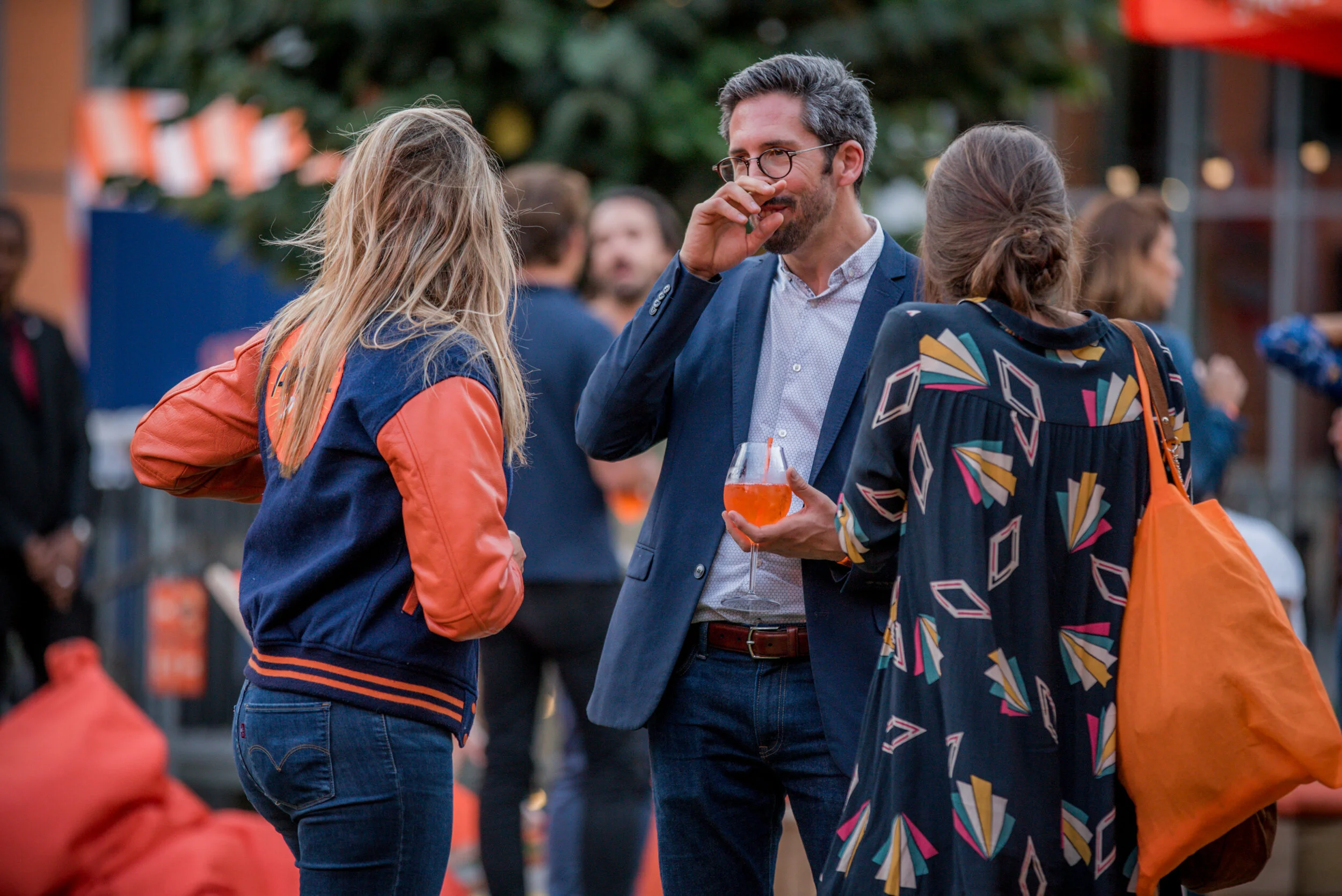 Groupe de trois personnes conversant lors d'un événement en extérieur, avec des décorations de Noël, une personne tenant un verre de cocktail orange, tous habillés de façon décontractée.
