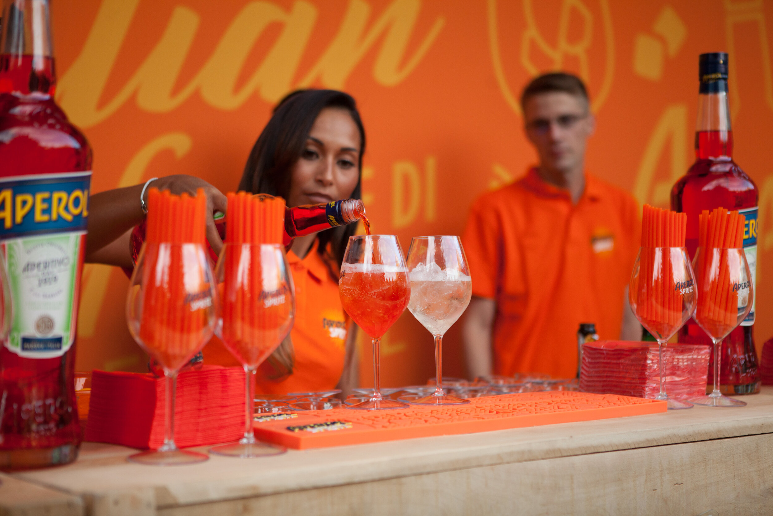 Deux personnes en uniforme orange préparent des cocktails avec des bouteilles Aperol, des verres contenant une boisson rouge et blanche, sur un bar orange.