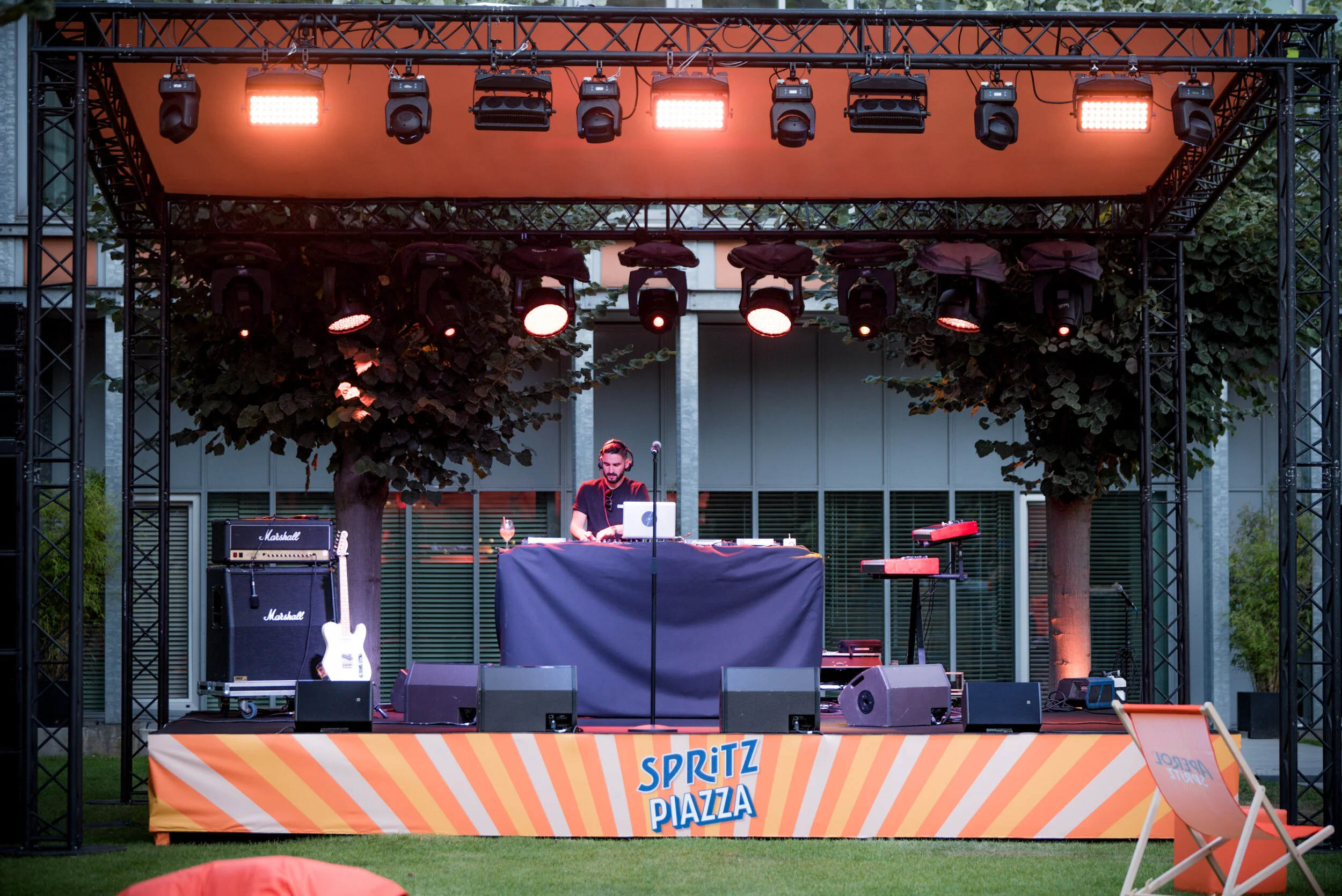 Un DJ joue sur une scène en plein air avec des lumières rouges, entouré d'équipements de musique, avec un panneau affichant 'Spritz Piazza' et des chaises pliantes orange devant la scène.