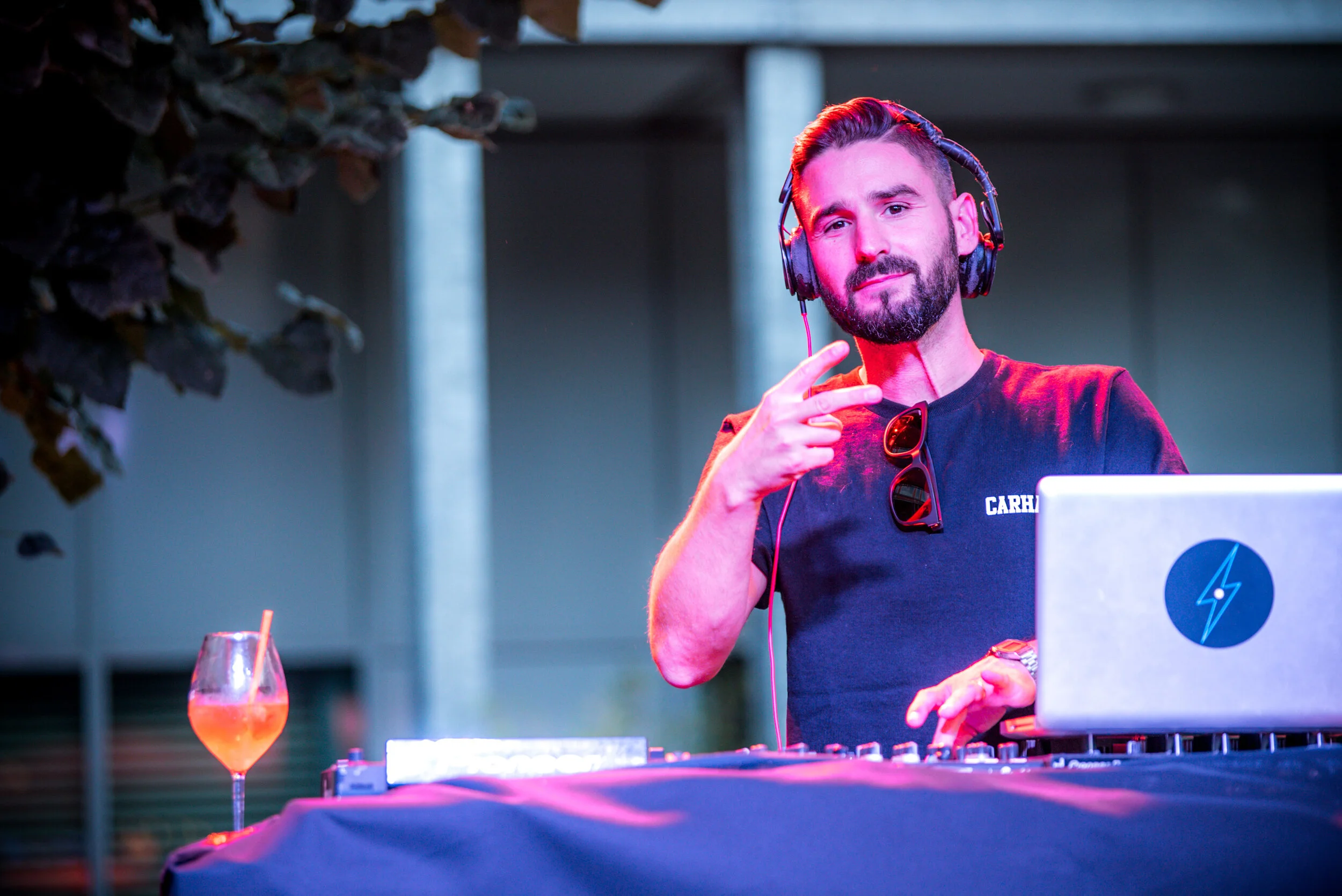 Un DJ souriant portant un t-shirt noir avec des lunettes de soleil accrochées, portant des écouteurs, se tient derrière une console de mixage avec un ordinateur portable à côté, pendant une soirée en extérieur, à côté d'un verre de cocktail orange av