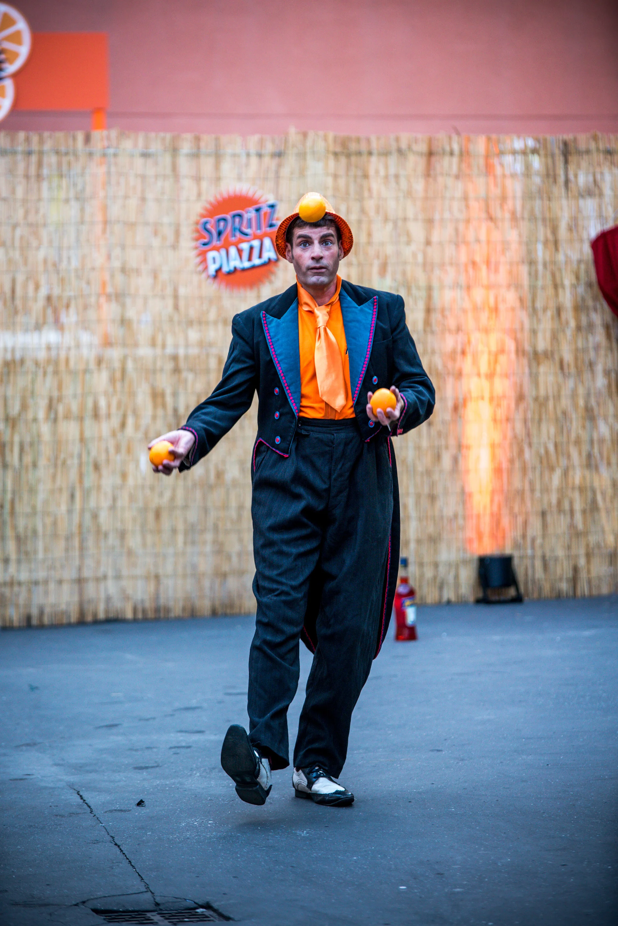 Un homme déguisé en clown jongle avec des balles orange sur scène, avec un fond en paillettes et un panneau "Spritz Piazza".