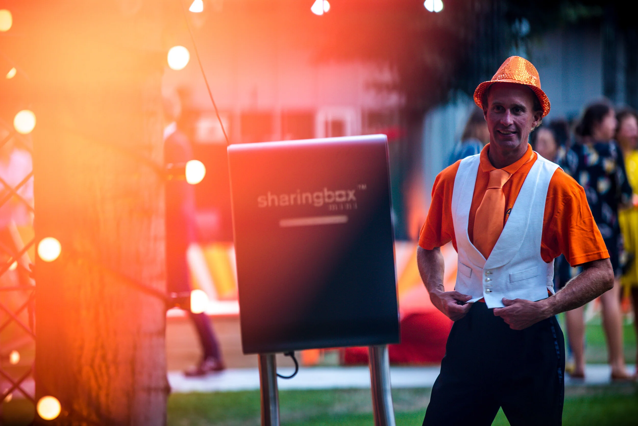 Un homme habillé en costume orange et blanc avec un chapeau orange, debout près d'un panneau d'information dans un événement en plein air, avec des personnes en arrière-plan.