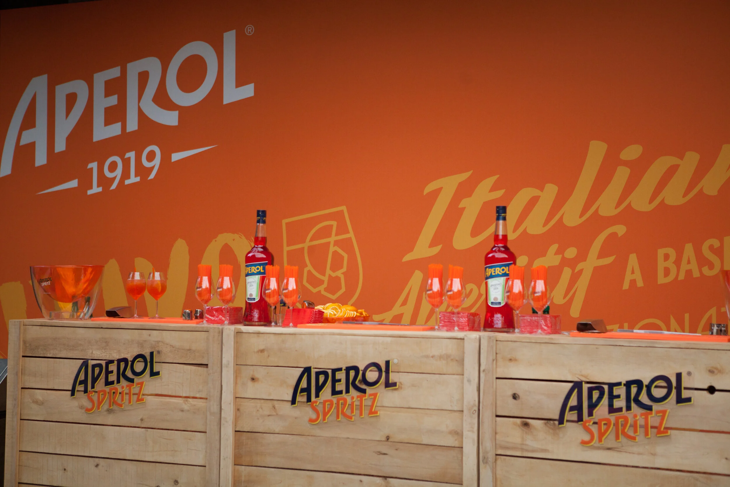Stand promotionnel d'Aperol Spritz avec bouteilles, verres et chips sur un fond orange avec le logo Aperol et la mention "1919".