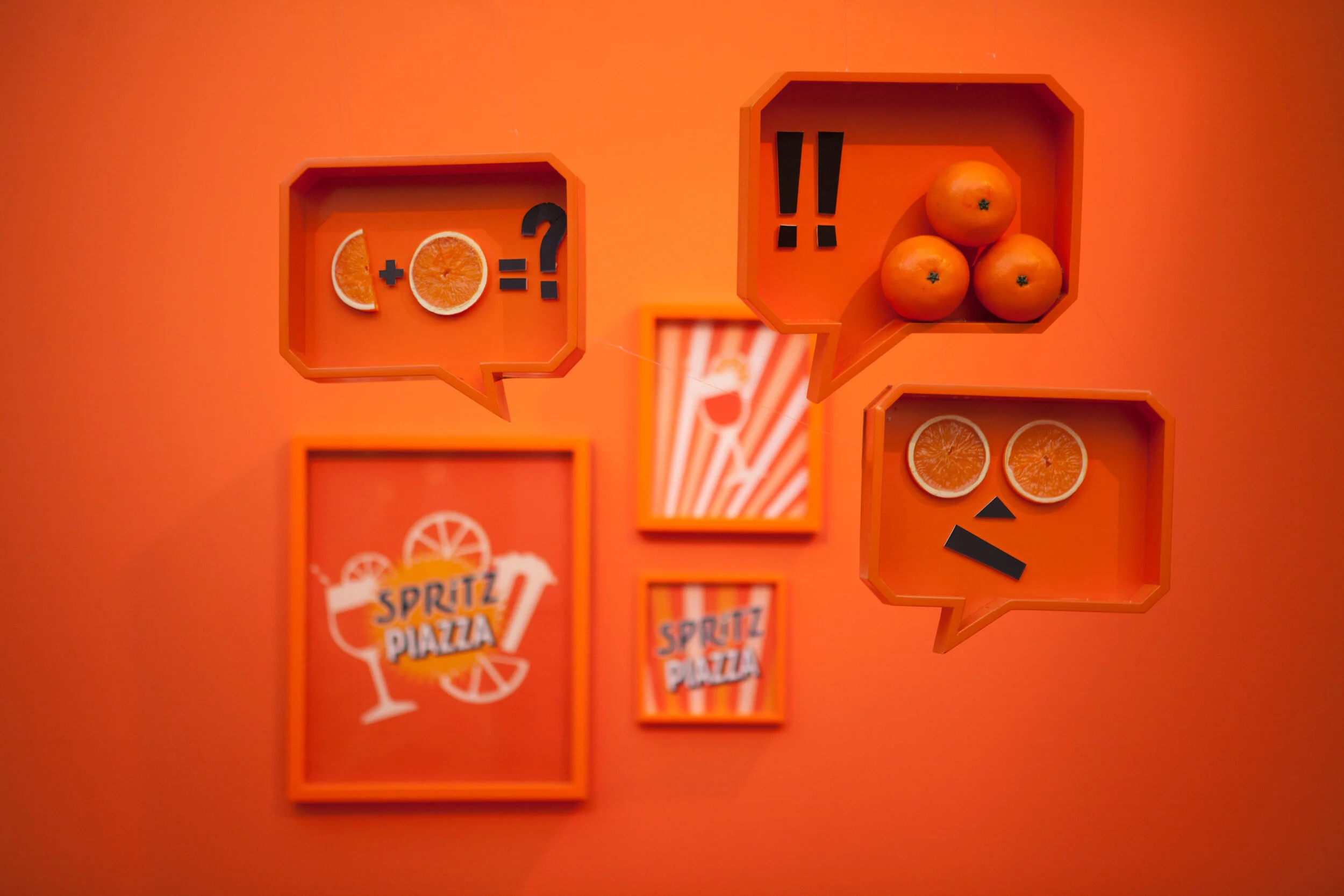 Décor orange avec boîtes de rangement, oranges, images de citrons, symbole de question, et le logo 'Spritz Piazza'.